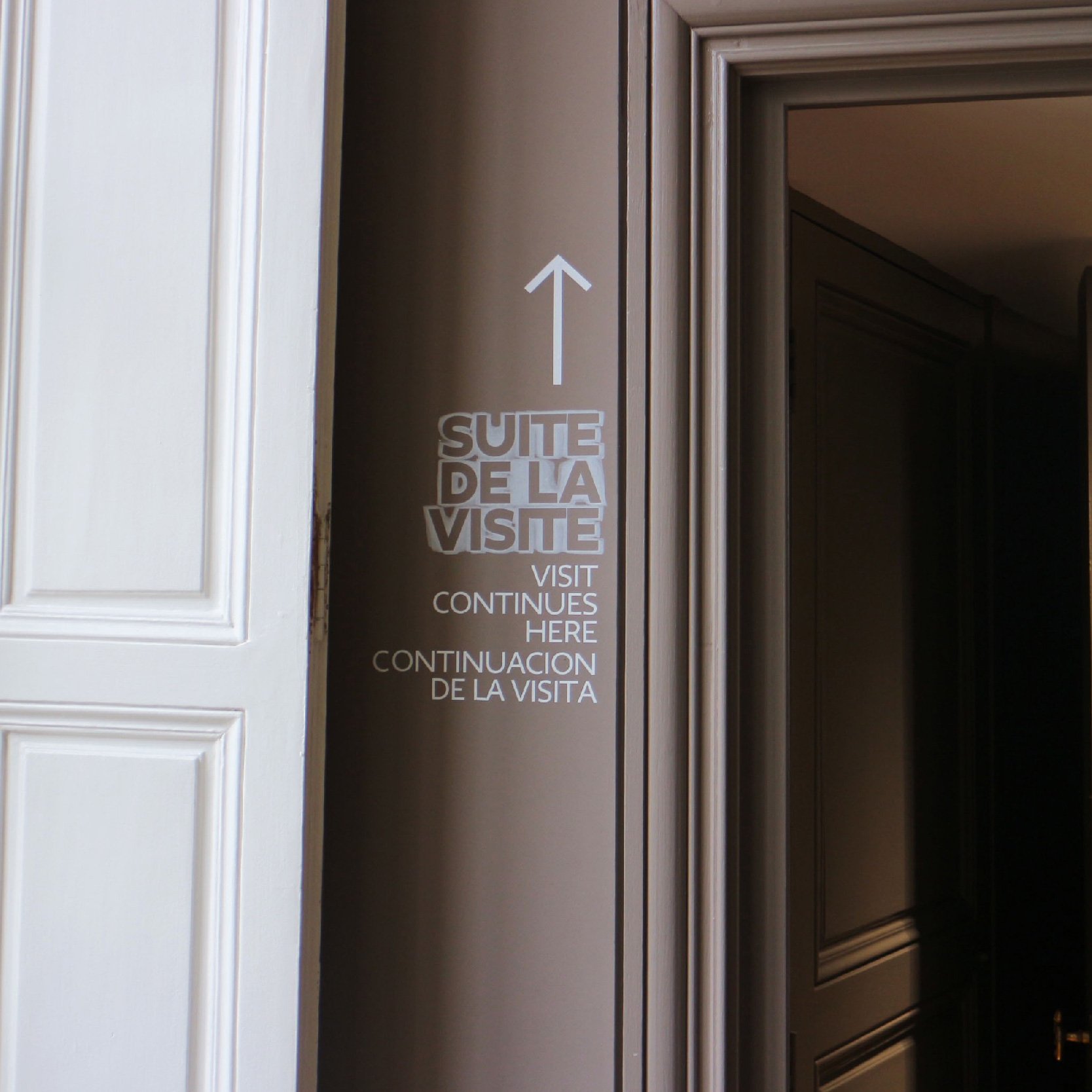 Signalétique « Suite de la visite » avec flèche sur une cloison du Musée Rodin, texte en trois langues et lettrage en relief
