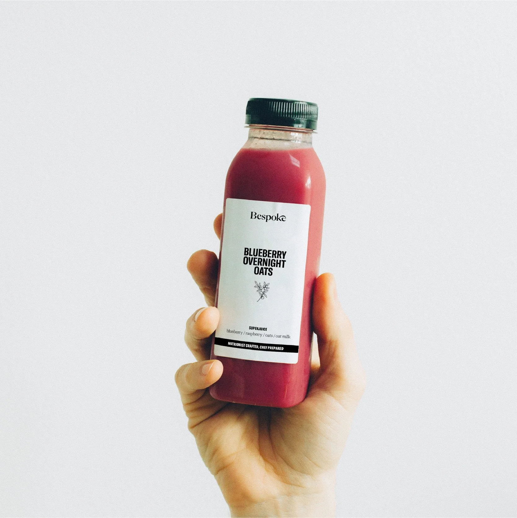Main tenant une bouteille de jus Bespoke « Blueberry overnight oats » à étiquette blanche, packaging de boisson