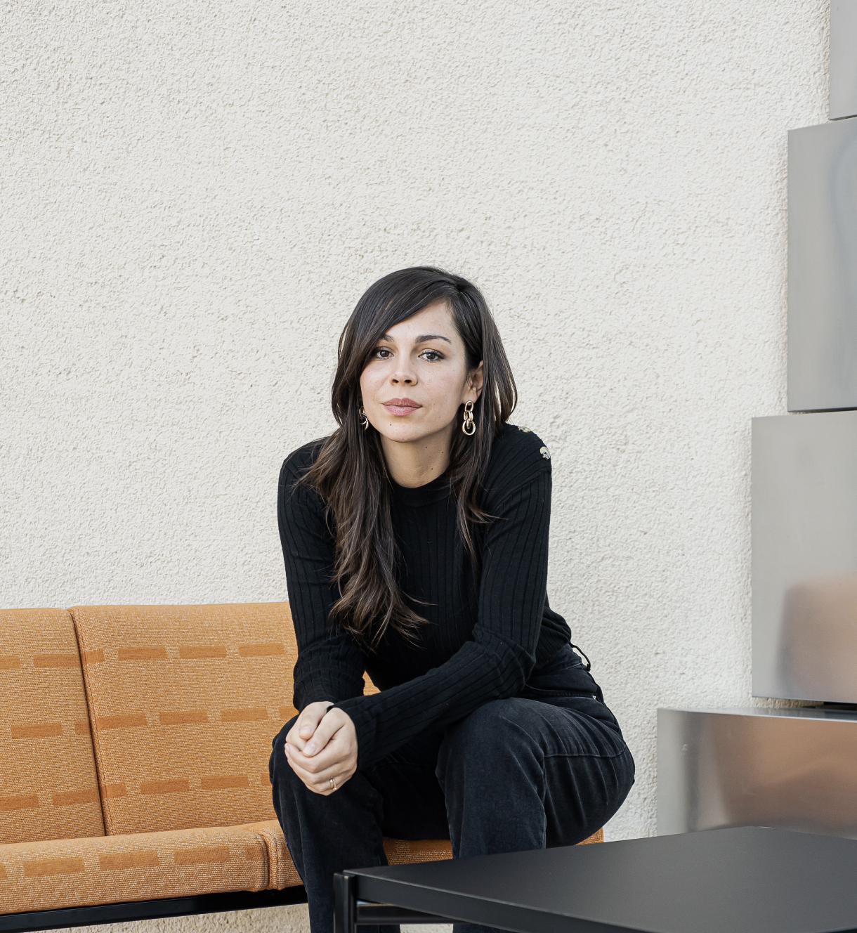 Une femme assise sur un canapé orange, portant un pull noir à rayures, avec des boucles d'oreilles en métal, devant un mur beige, dans un endroit moderne.
