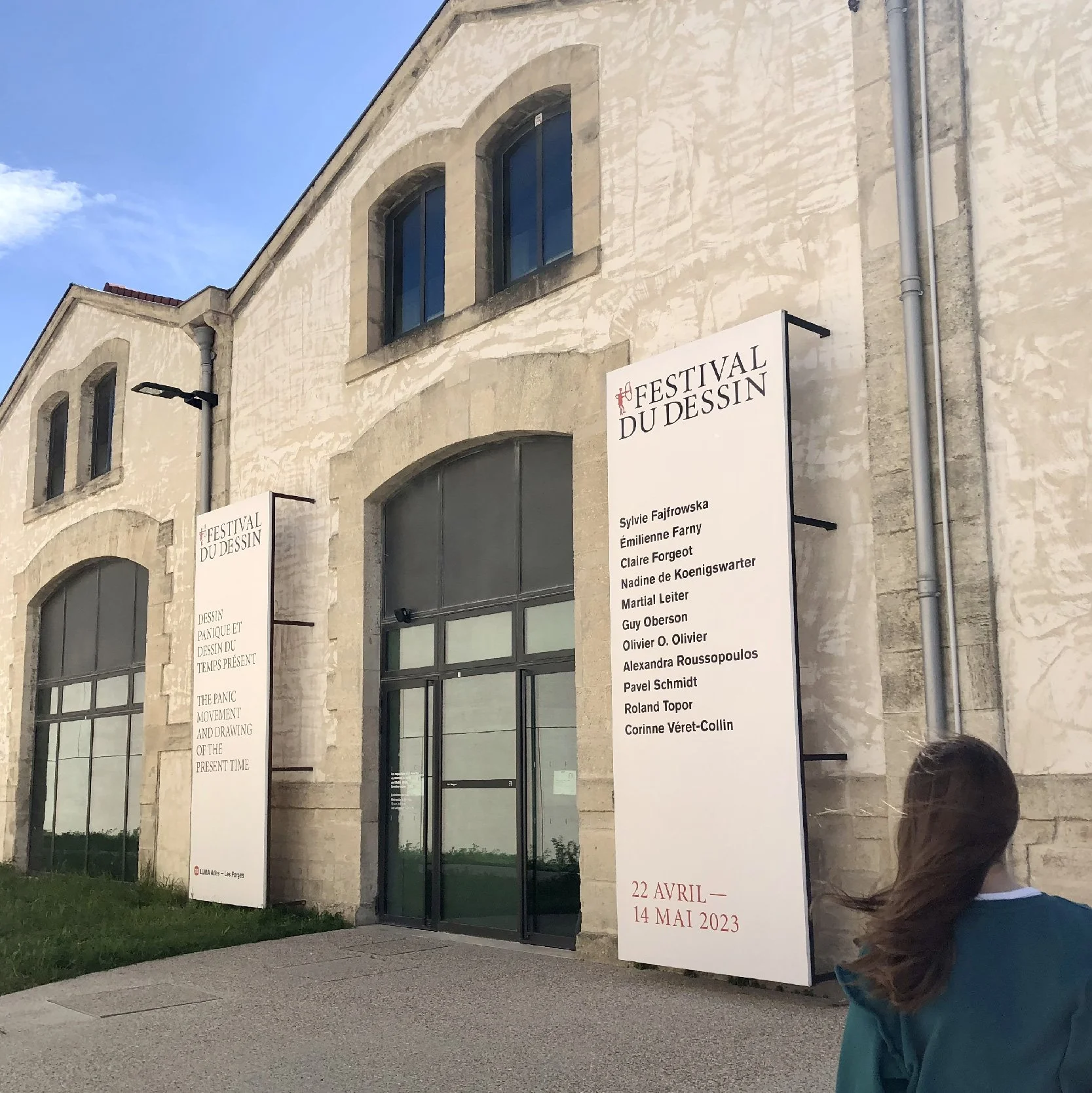 Grands panneaux verticaux devant un bâtiment en pierre, liste des artistes invités, signalétique d'entrée du Festival du dessin