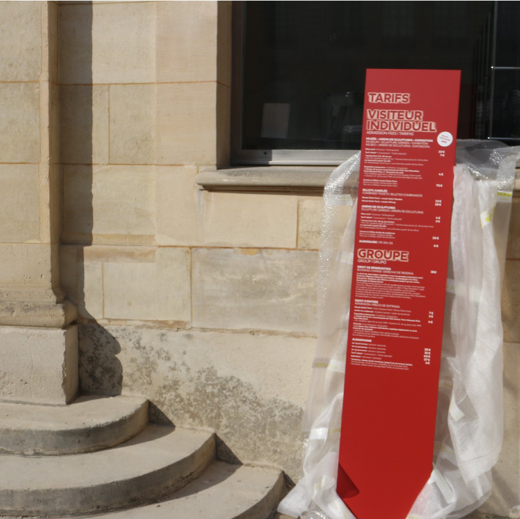 Totem rouge des tarifs « Visiteur individuel » et « Groupe » devant la façade en pierre du Musée Rodin