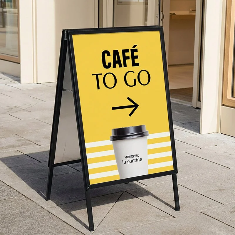 Panneau publicitaire indiquant "Café To Go" avec une flèche pointant vers la droite, une tasse à café blanche avec un couvercle noir et le texte "MONOPRIX la cantine".