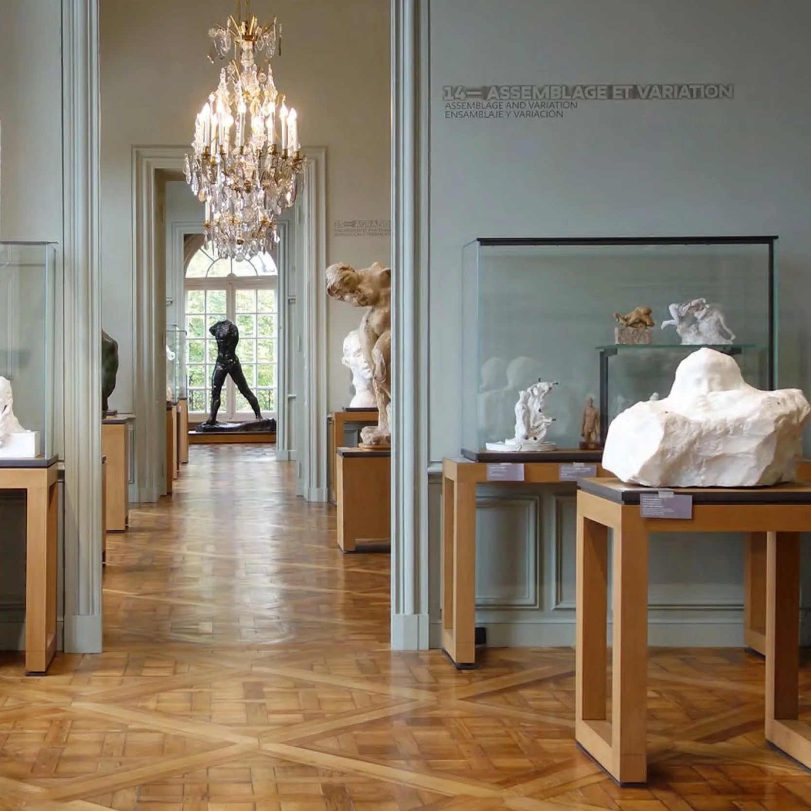 Exposition de sculptures dans une salle d'un musée ou une galerie d'art. Plusieurs statues en pierre et en bronze exposées sur des socles en bois, un lustre suspendu au plafond, fenêtres laissant entrer la lumière naturelle.