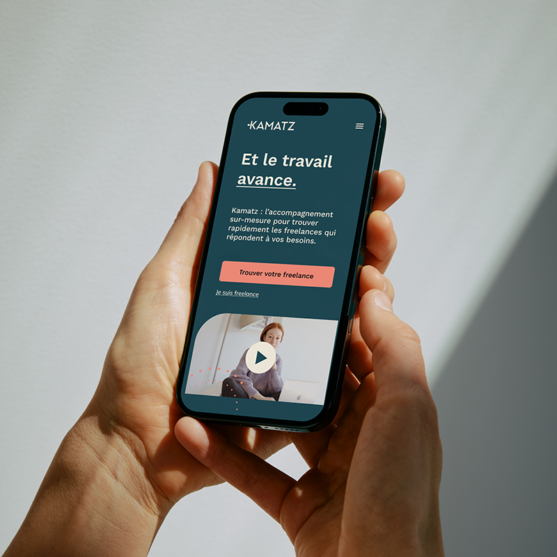 Une personne tient un smartphone affichant une application appelée 'KAMATZ' qui propose un accompagnement personnalisé pour trouver des freelances rapidement, avec un bouton pour trouver sa freelance et une vidéo en lecture automatique.