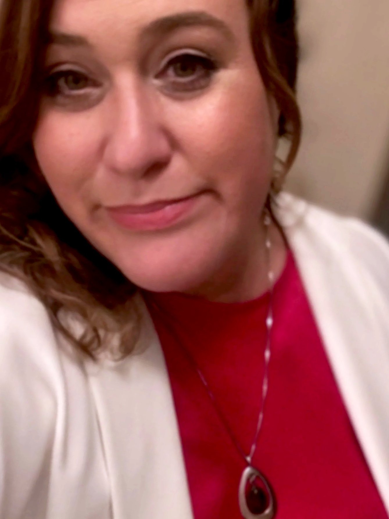 Close-up of a woman with light brown, wavy hair, wearing a red top with a matching jewelry, and a light-colored jacket.