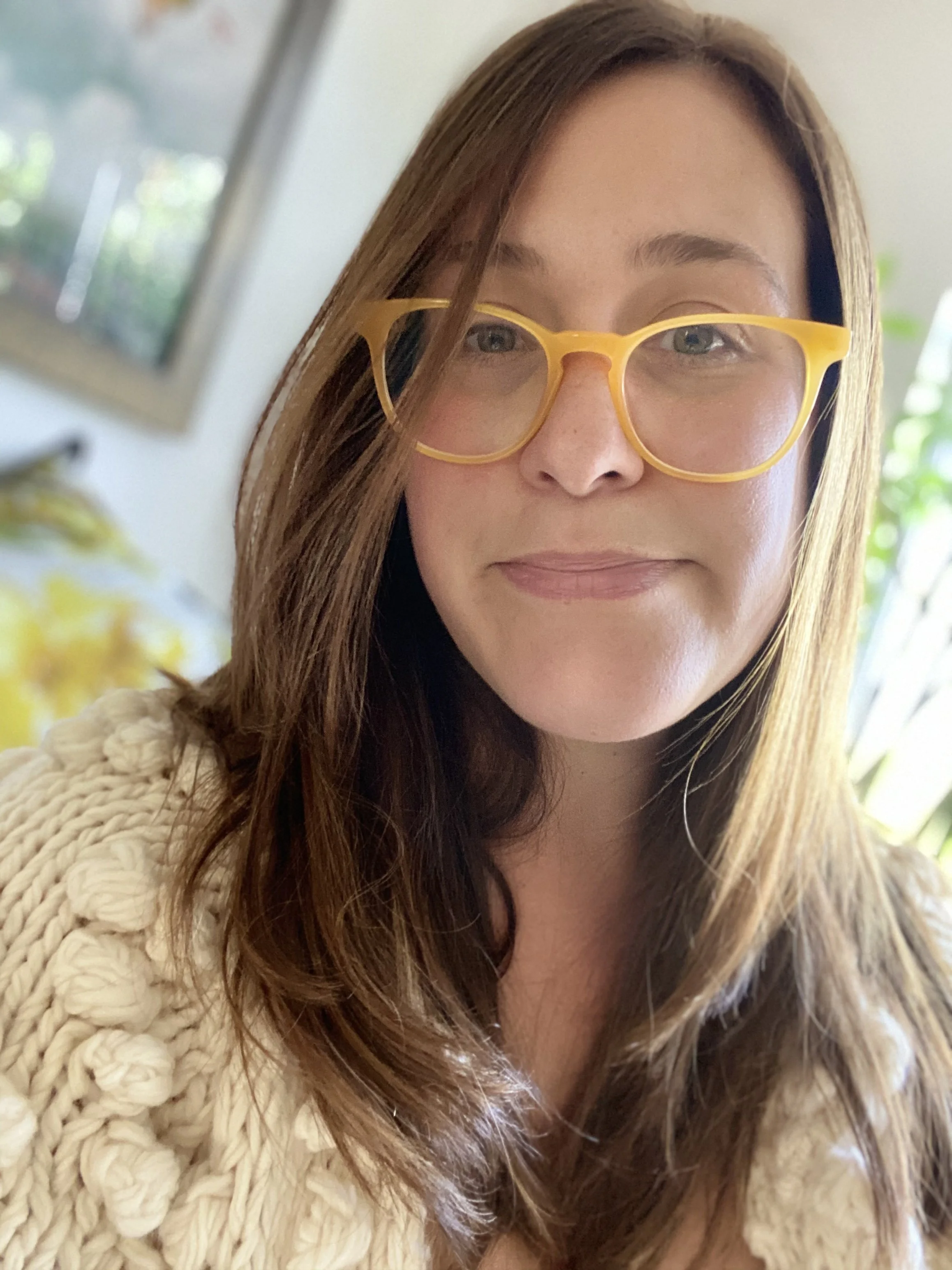 Close-up of a woman with long brown hair wearing yellow glasses and a cream-colored knitted sweater.