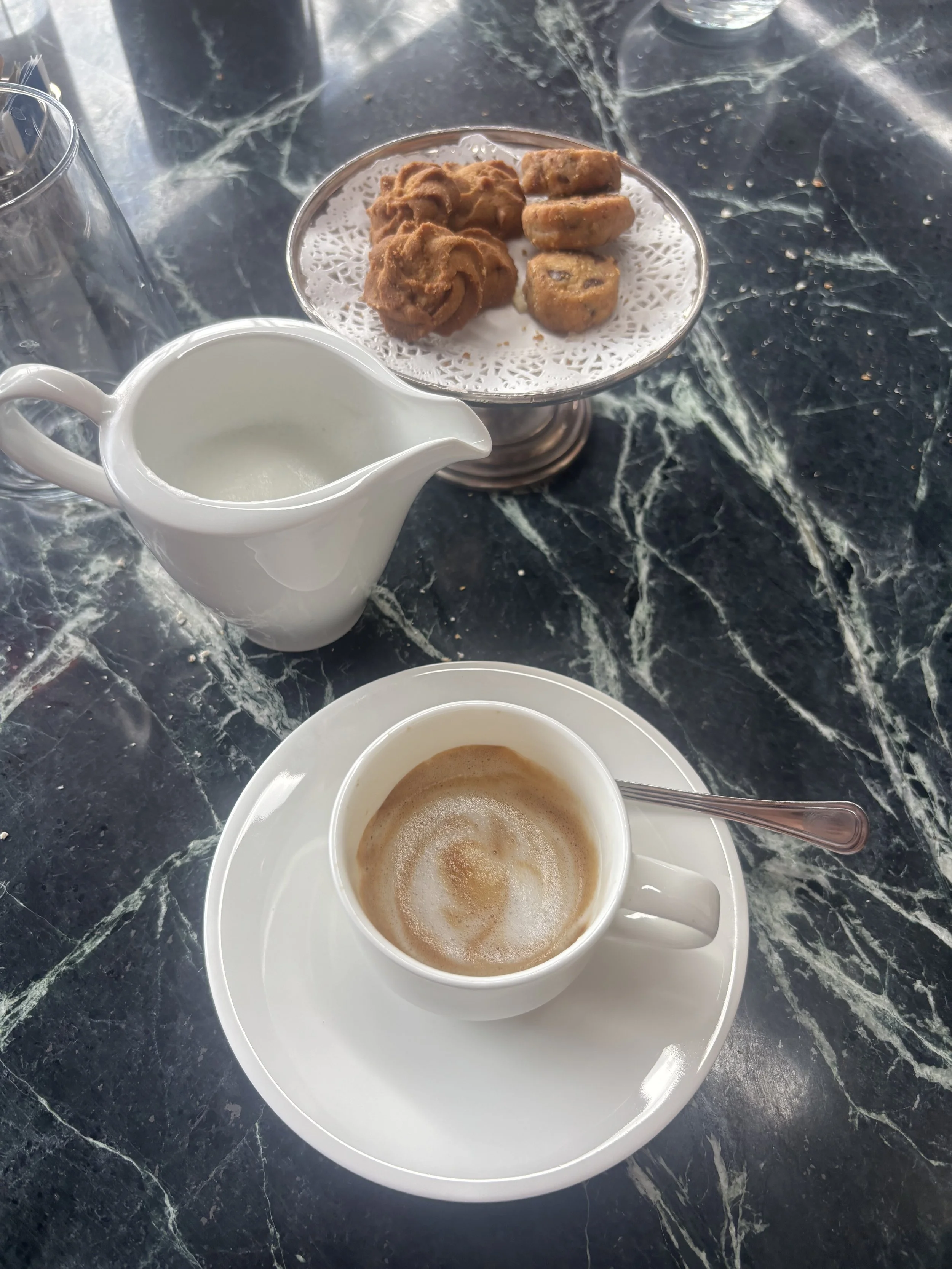 A white cup of coffee with foam art, a small white pitcher of milk, and a plate of cookies on a marble table.