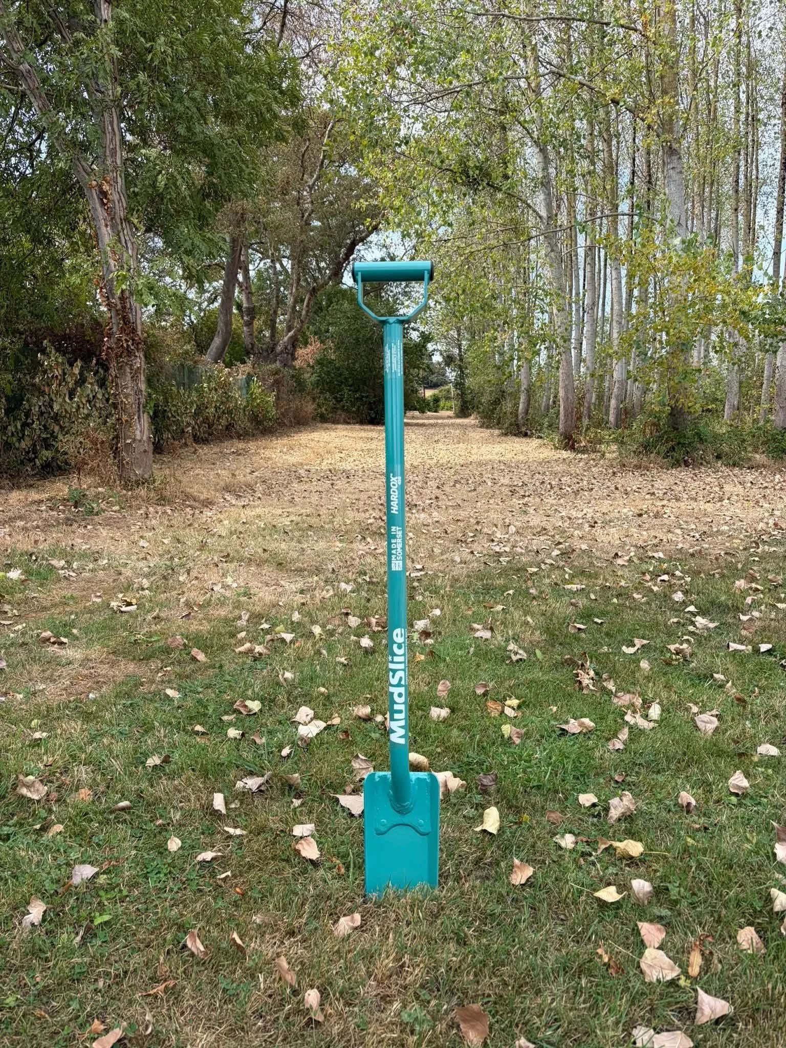MudSlice Trench Rat Drainage spade standing in a grassy park area with trees and fallen leaves surrounding it.