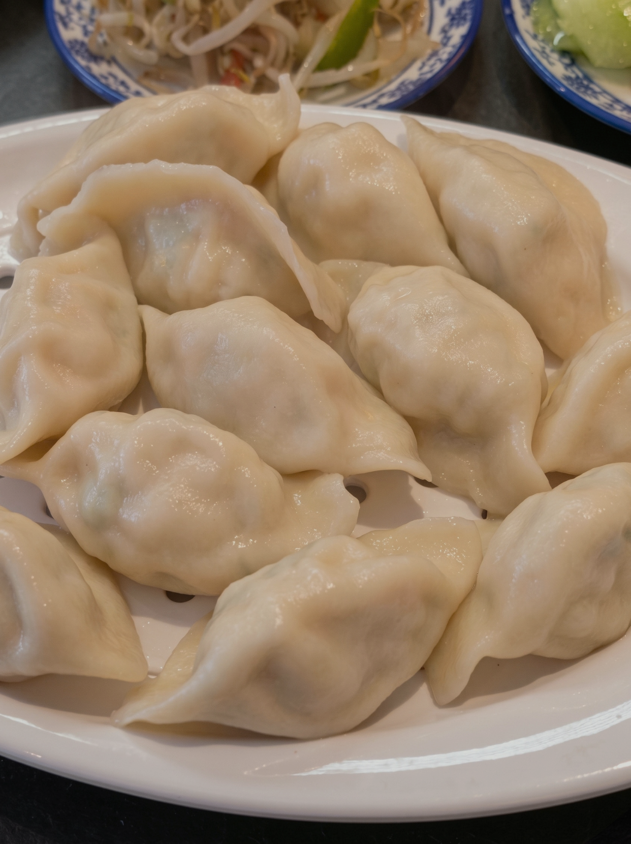 Close-up of steamed dumplings served on a white plate with side dishes of noodles and lettuce in blue-patterned bowls in the background.
