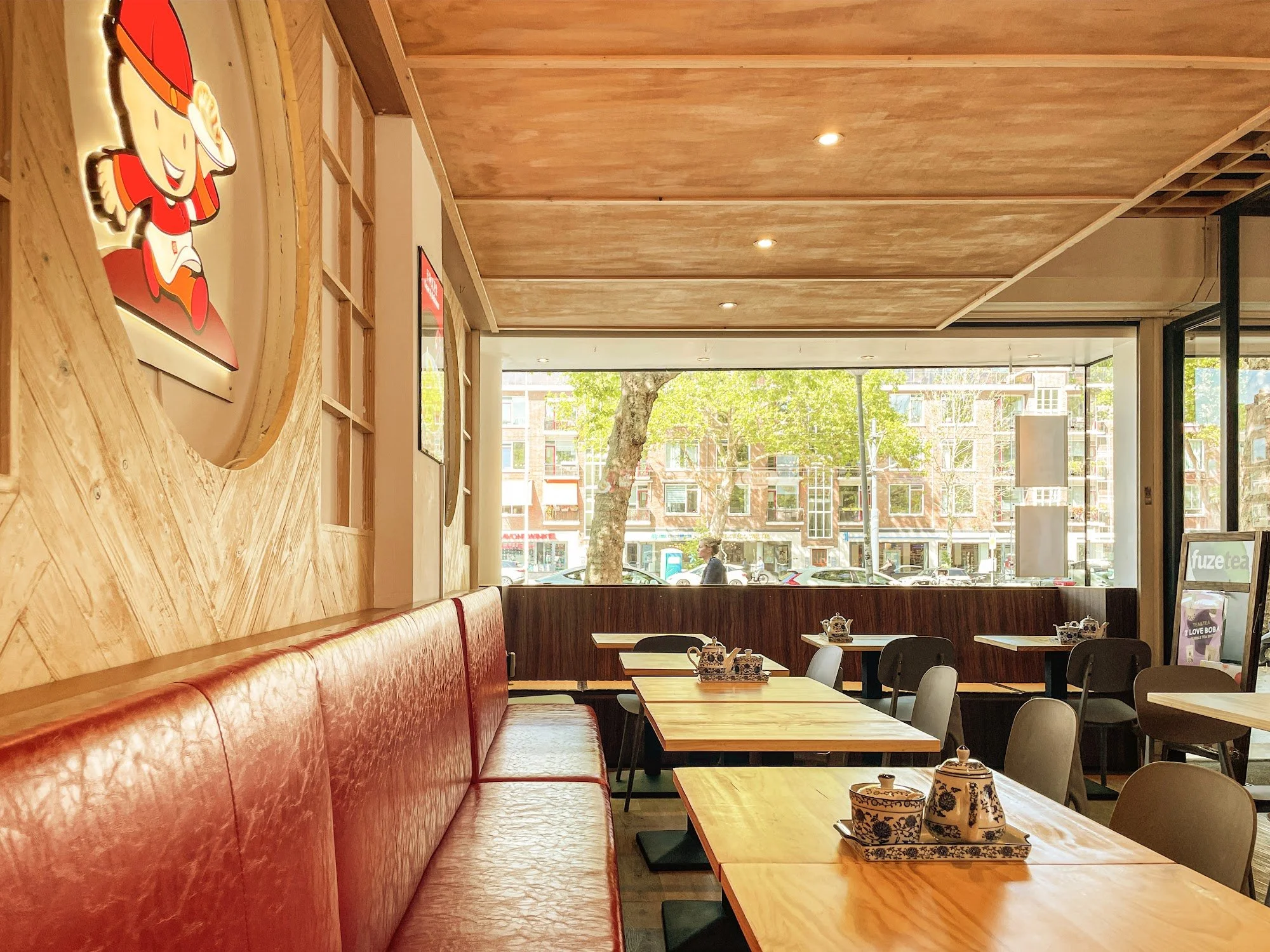 Interior of a cozy cafe with wooden ceiling panels, large windows, and a view of the street with trees and buildings outside. There are tables set with teapots and cups, and a red leather bench along the wall.