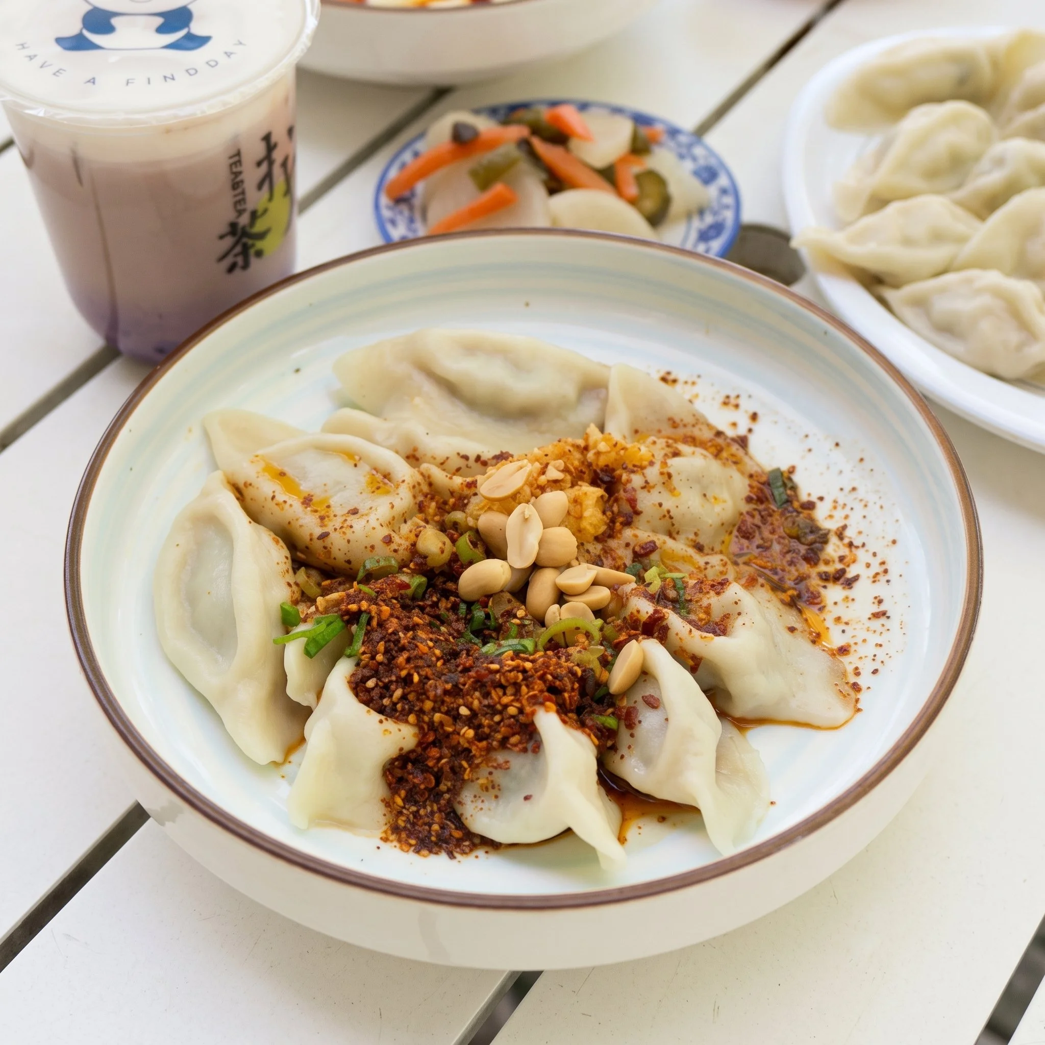 A bowl of Asian dumplings garnished with crushed peanuts, chopped green onions, and chili flakes. In the background, there is a small dish of pickled vegetables and a cup of bubble tea with a straw.