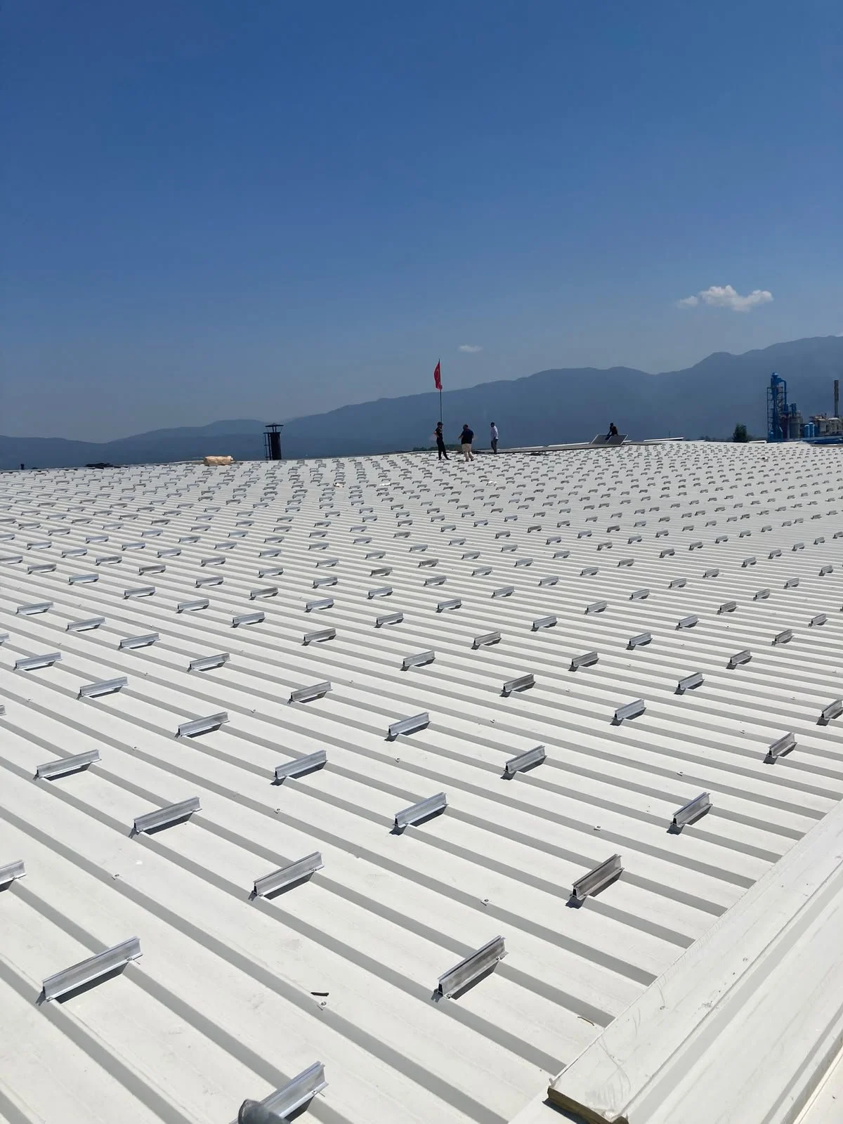 Gümüş renkli enerji panelleriyle kaplı büyük bir çatı, açık mavi gökyüzü ve arka planda dağlar, çatıda 3 kişi ve Türk Bayrağı bulunuyor.