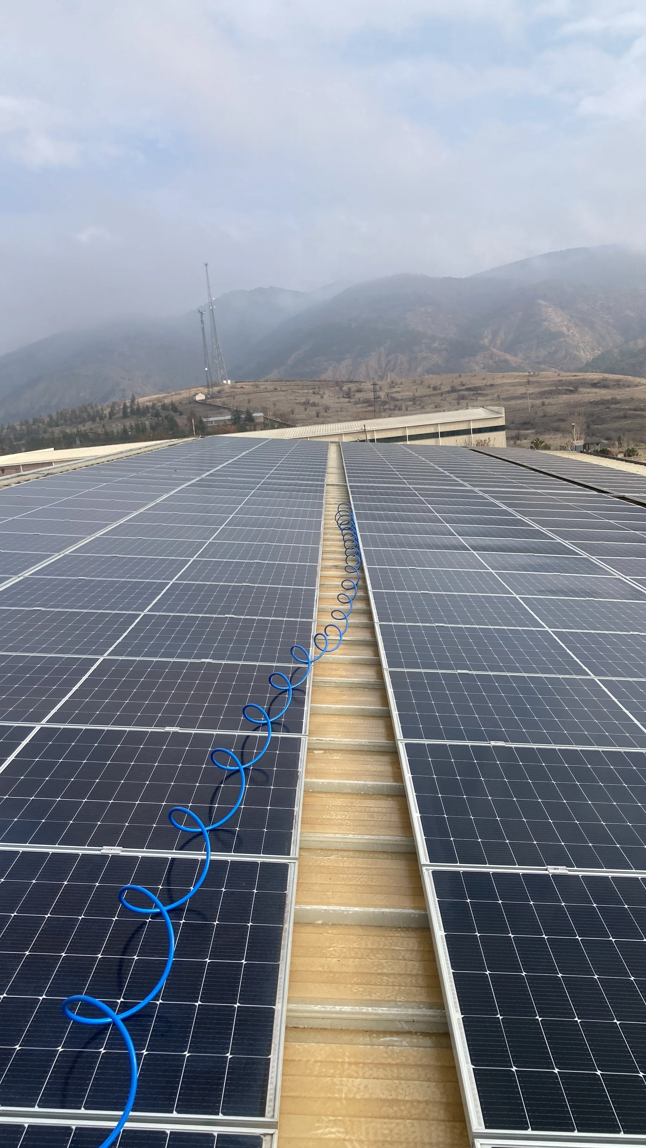 Güneş enerjisi panelleriyle donatılmış bir çatı, arka planda dağlar ve gökyüzü görünmekte.