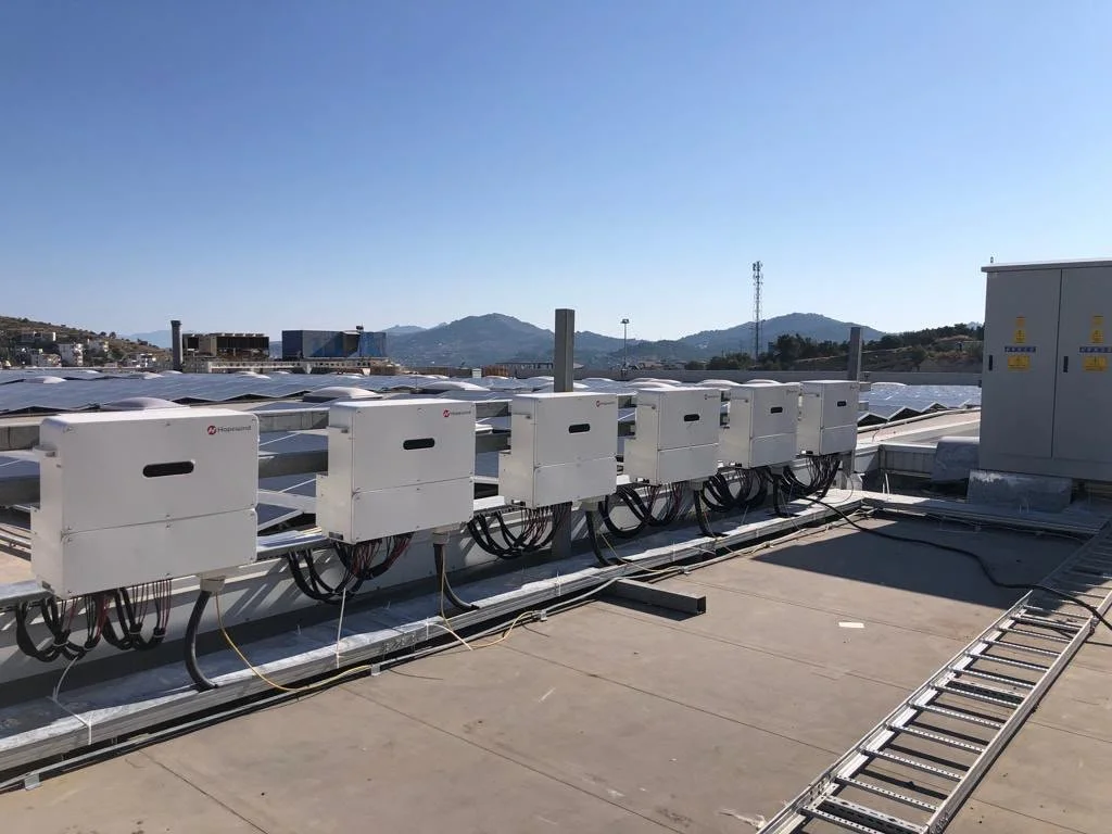 Güneş enerjisi panelleri üzerinde kurulu inverter ve elektrik kutuları, açık hava ortamında, şehir manzarası ve dağlar görünümünde.