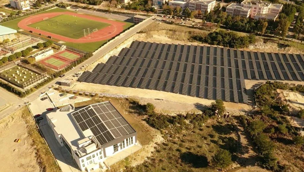 Güneş panelleriyle kaplı büyük bir bina, yanında güneş enerjisi panelleriyle dolu geniş bir alan, okullar ve spor sahaları çevresinde, ağaçlıklı bölgeyle çevrili.