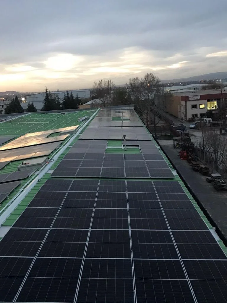Büyük bir binanın çatısında güneş panelleri ve güneş enerji sistemleri yer alıyor, arka planda bulutlu gökyüzü ve şehir manzarası görülüyor.