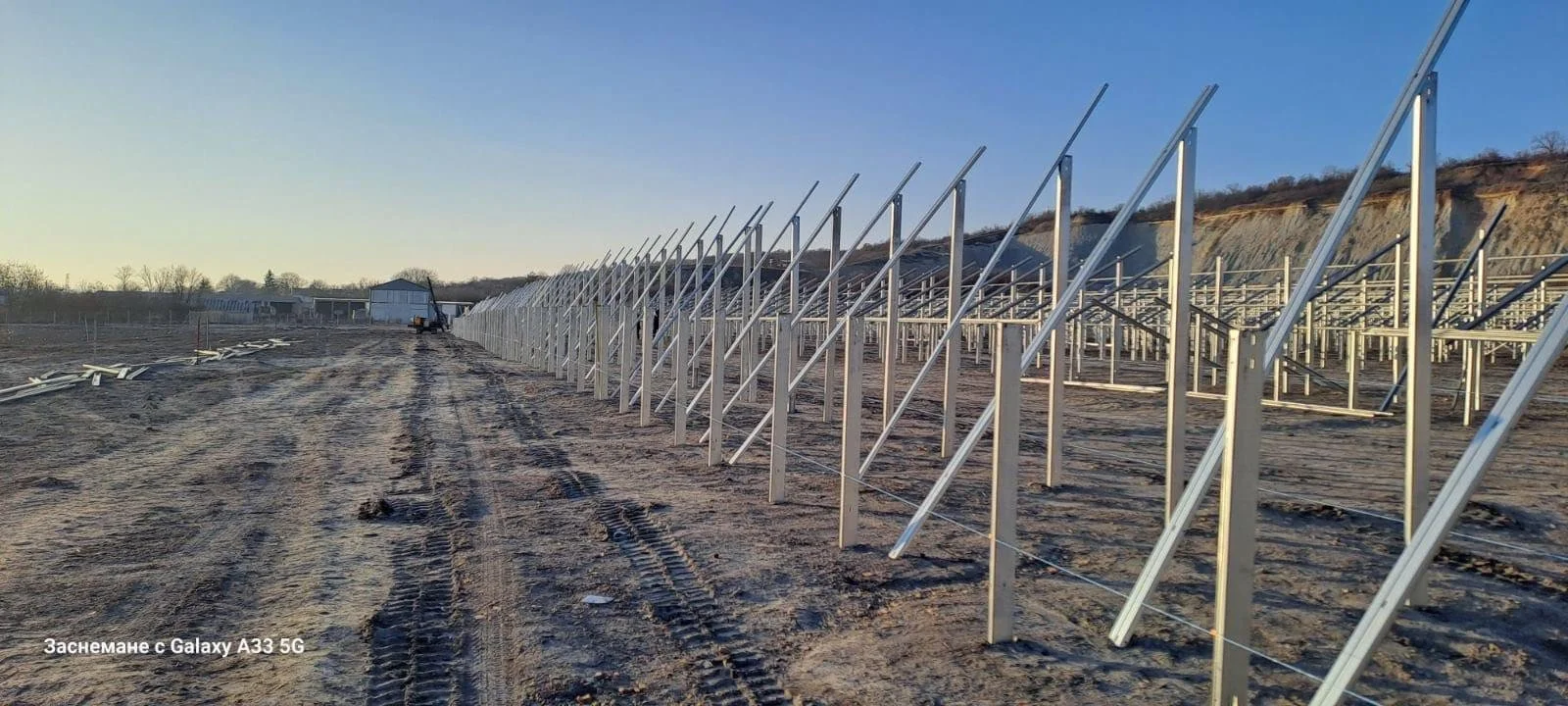 Güneş enerjisi panelleri kurulumu sırasında metal çerçevelerin montajı yapılan açık alanda boş ve toprak zemin. Panellerin çerçeve yapısı yerleştiriliyor, teçhizat ve araçlar uzaklarda görünüyor.