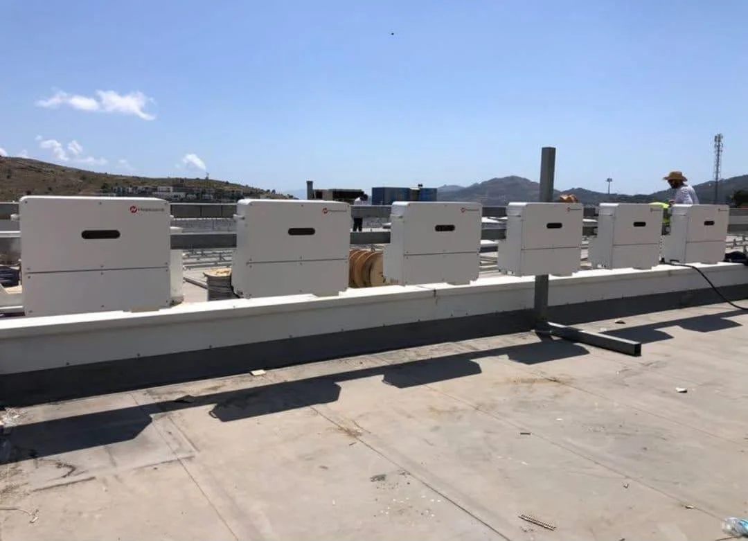 Çatıdaki beyaz elektrik ekipmanları ve kabloları, gökyüzü ve uzak dağlar, bir kişi şapka ve güvenlik yeleği ile çalışıyor.