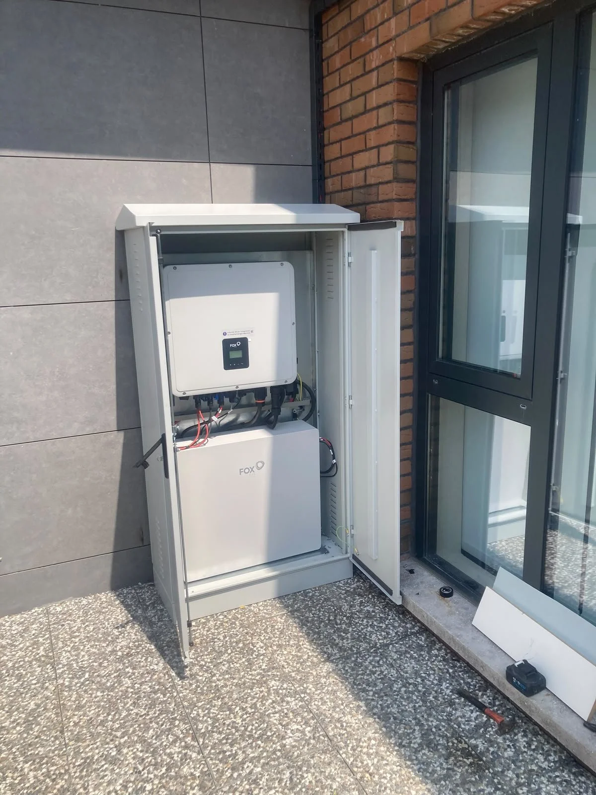 Dış mekanda duvara monte edilmiş elektrikli cihaz, kapak açılmış açılımda, iç kısmında çeşitli bağlantılar ve bir dijital ekran var. Etrafında mermer ve tuğla duvarlar, yerde kazıma ve araçlar bulunuyor.