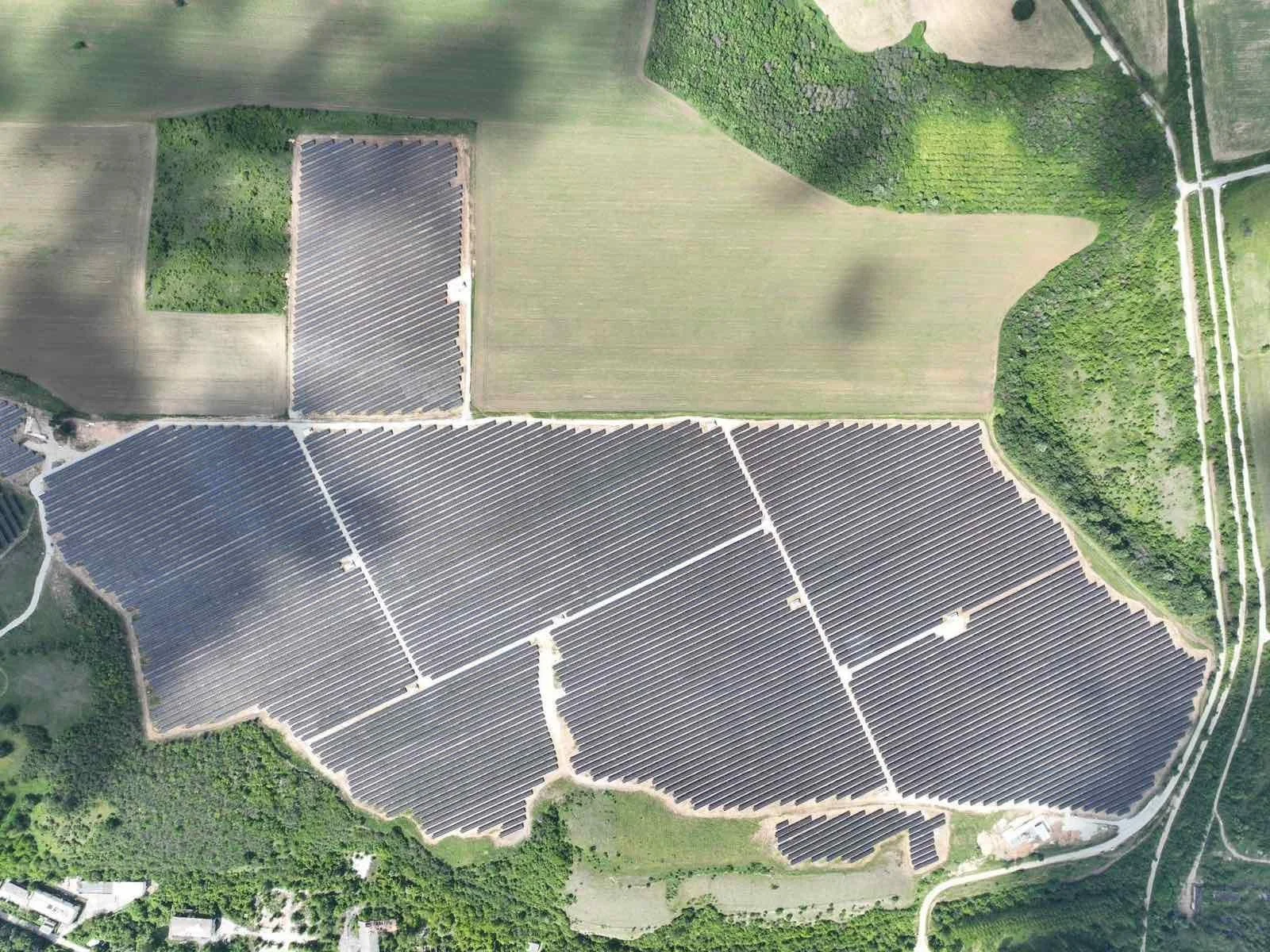 Geniş bir güneş enerjisi panelleri tarlası, çevresinde ağaçlık ve açık alanlar bulunan drone ile çekilmiş bir hava fotoğrafı.
