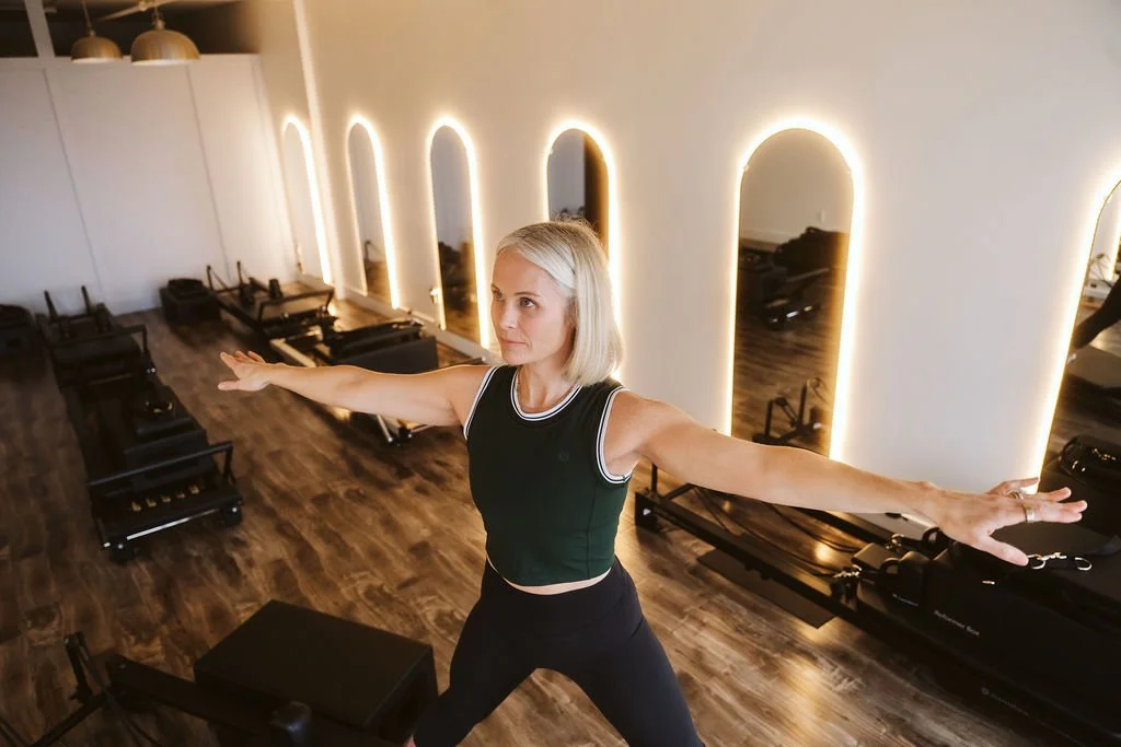 Woman with blonde hair stretching arms in a fitness studio with mirrors and Pilates reformers.