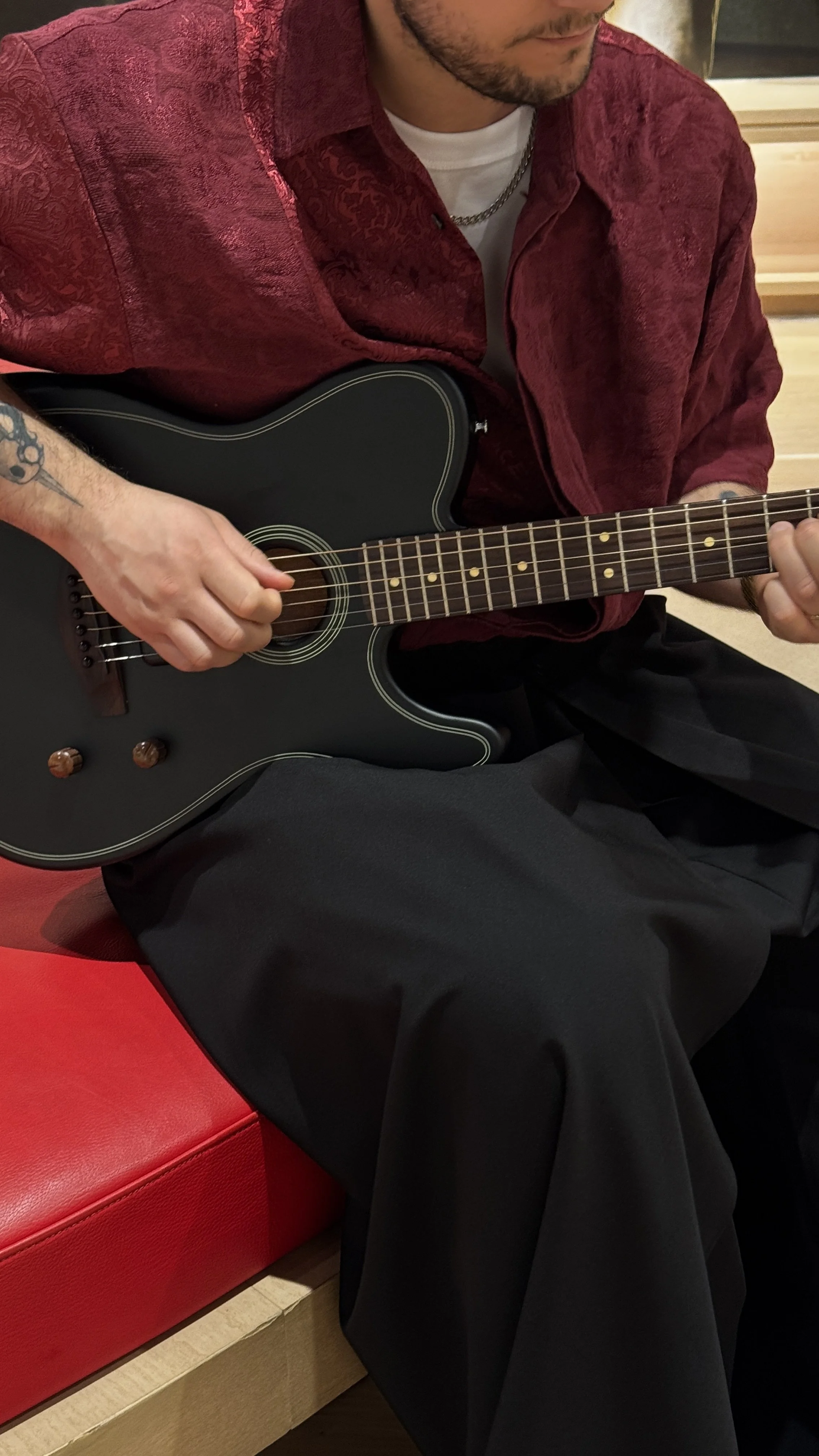 Person som spiller akustisk gitar, sittende på en rød sofa