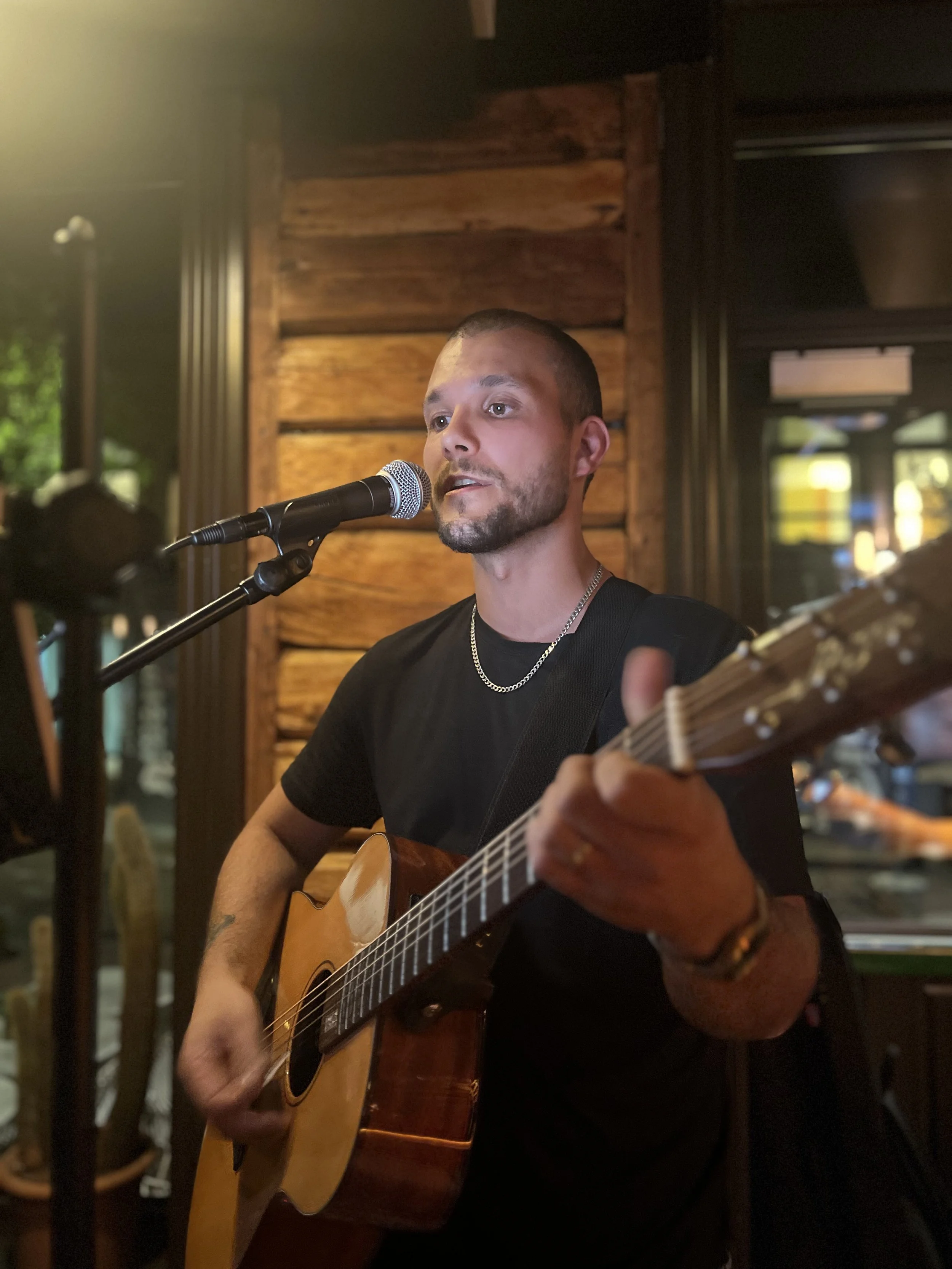 En mann spiller akustisk gitar og synger inn i en mikrofon i et innendørs miljø, med trevegger i bakgrunnen.