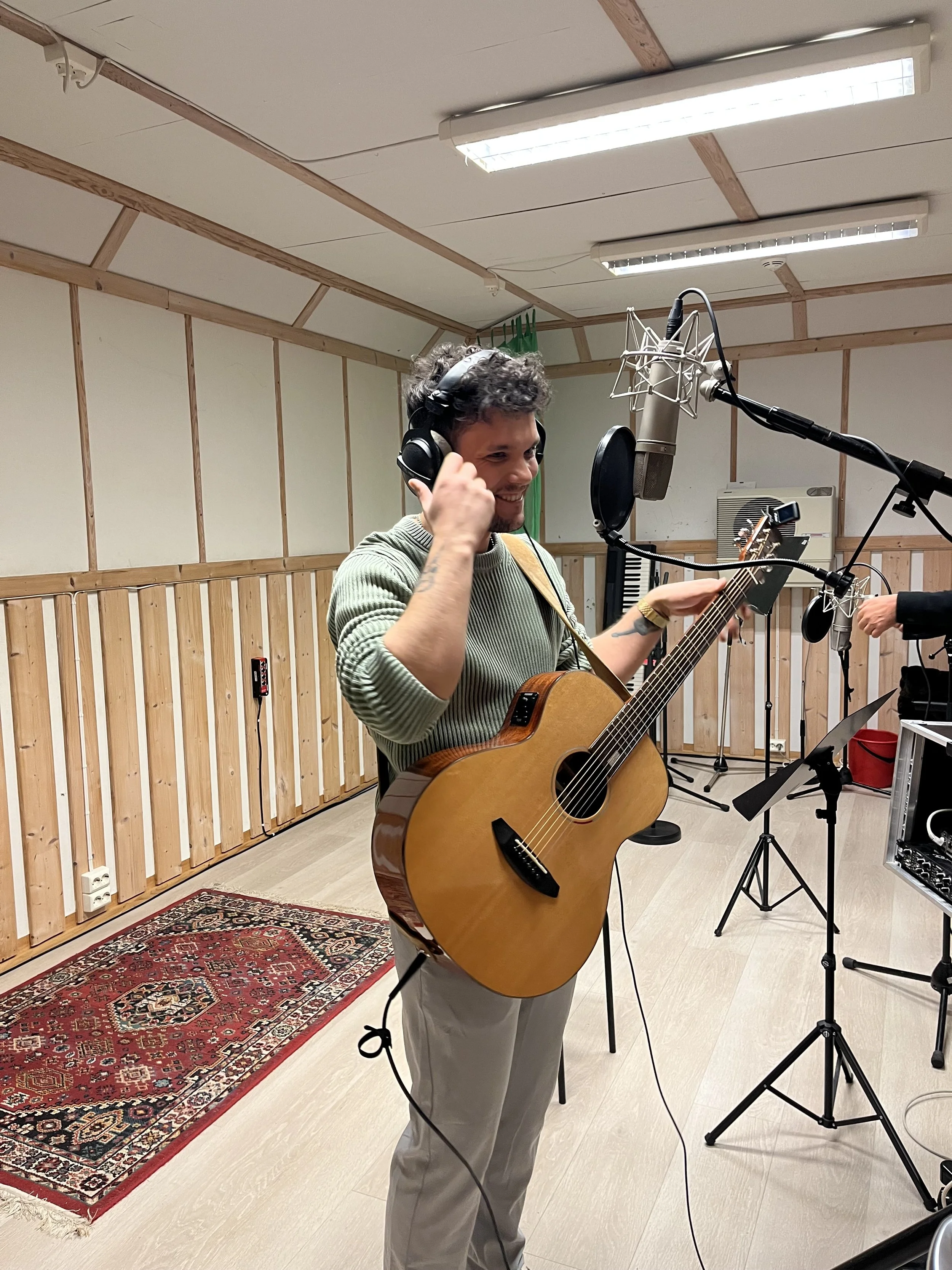 En mann med pent oppsatt hår spiller gitar i et studio med lydutstyr, mikrofoner og musikkutstyr, omgitt av trebjelker og lyddempende paneler.