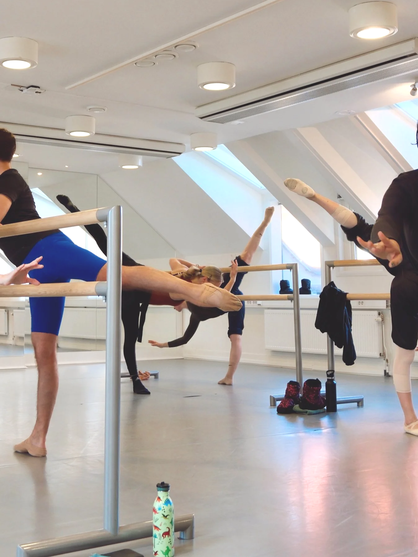 Kvinnor och män som utövar yogapositioner i en inomhussal, med barfota på guldfärgat golv, spegelväggar och takfönster. Linn Bergh , Balettcentrum Stockholm