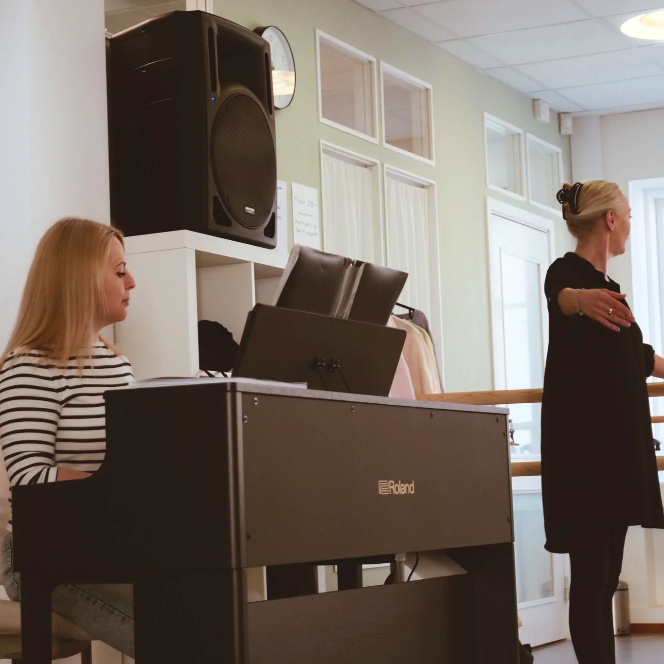 Två kvinnor, en som spelar piano och en annan som står och sjunger eller talar i ett rum med stora fönster. Anna Valev