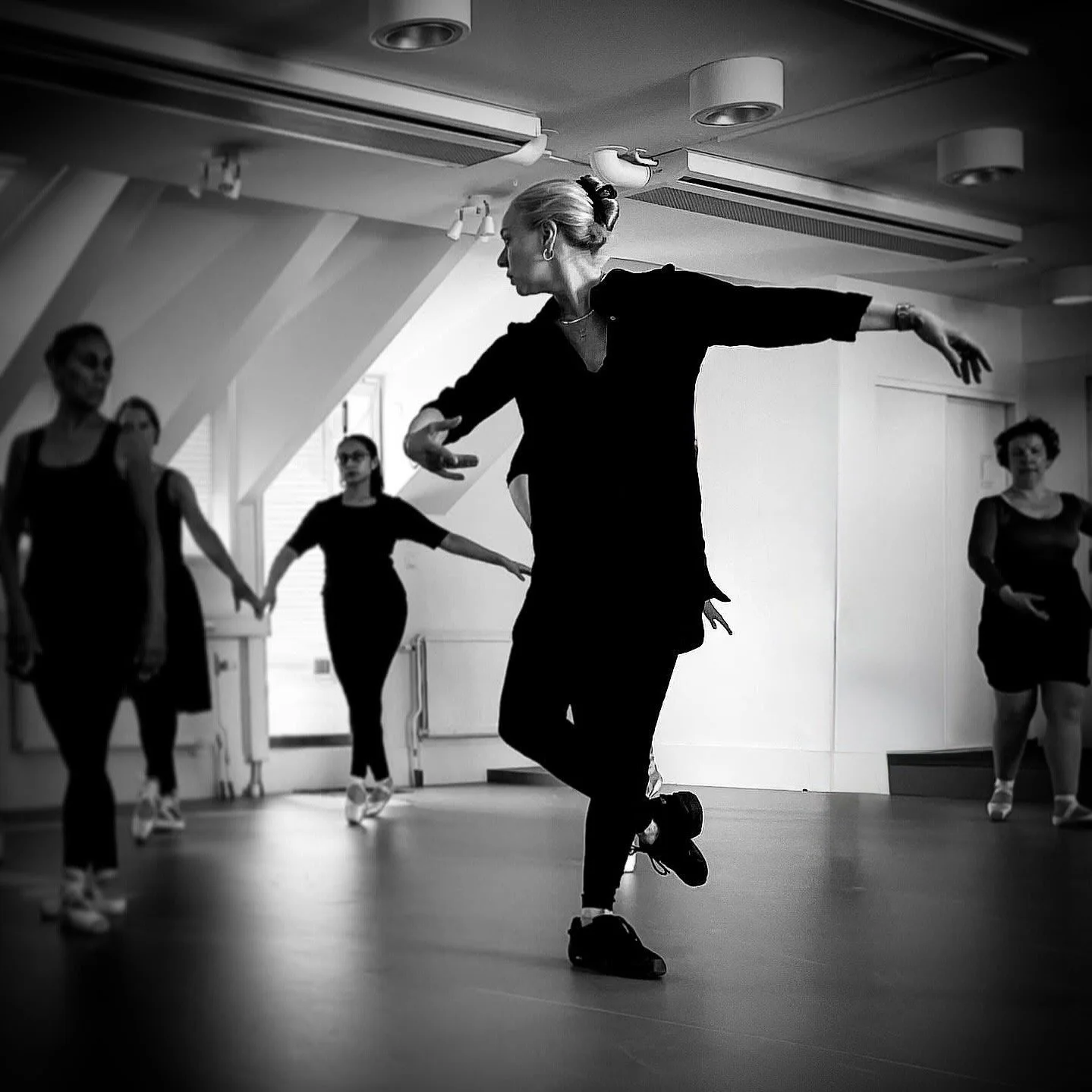 Anna Valev undervisar balett på Balettcentrum Stockholm. En grupp kvinnor som dansar i en studio, en kvinna i förgrunden dansar med utsträckta armar och ett lätt knäböjande steg, medan de andra står i bakgrunden.