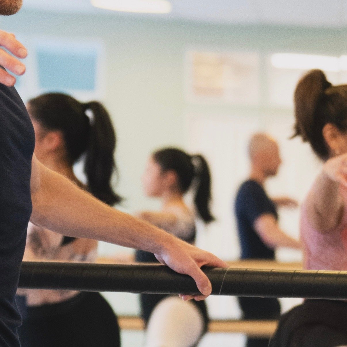 Person som håller i en balettstång i en dansstudio med flera personer som övar balletta i bakgrunden. Balettcentrum Stockholm