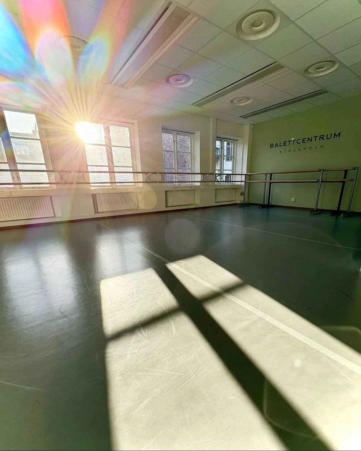 En tom balettsal med stora fönster som släpper in solljus, vägg med texten 'Balettcentrum Stockholm', grannd liknande golvytor och speglar längs väggarna. Balettcentrum Stockholm