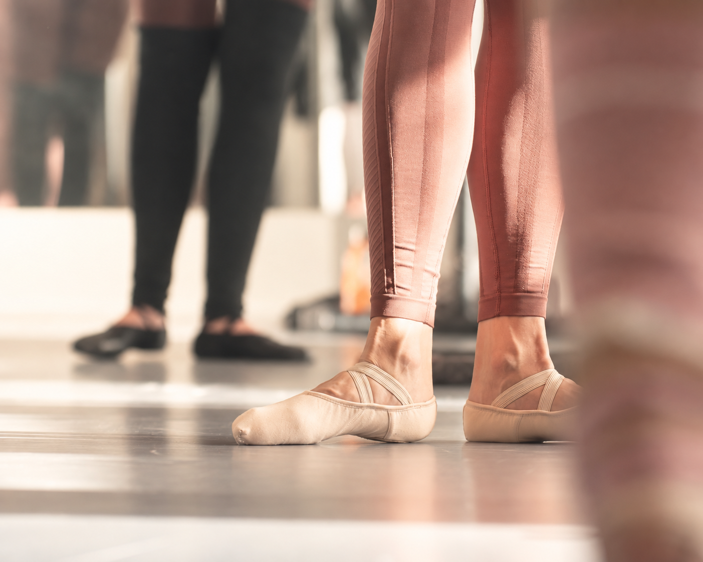 Närbild av en balettdansares fötter i rosa dansskor och rosa byxor, med andra personer i bakgrunden på en dansstudio. Balettcentrum Stockholm
