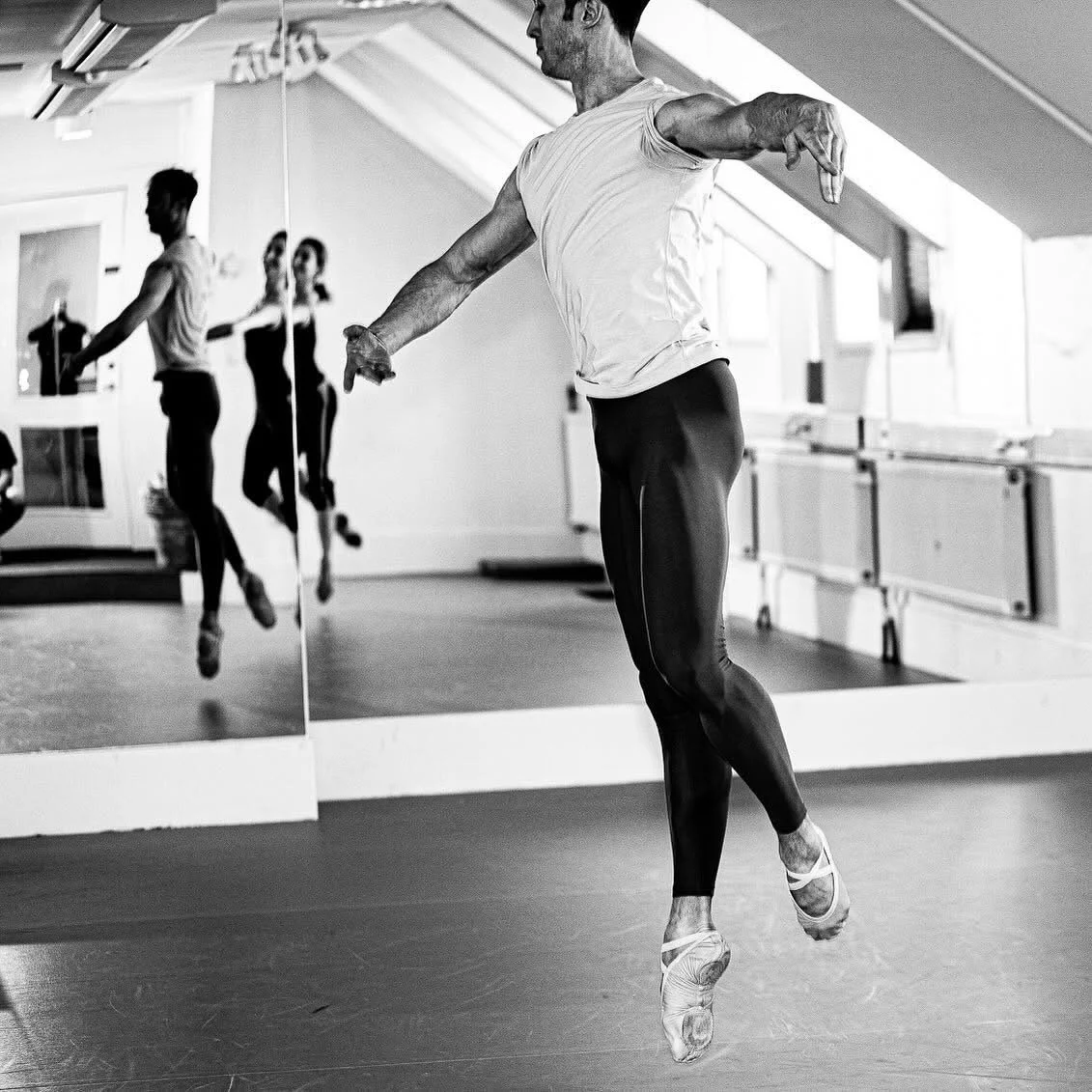 Bilden visar en man som tränar balett i en dansstudio med speglar. Mannen utför en piruett i balettkläder och balettskor, medan två andra personer syns i speglarna i bakgrunden. Daniel Goldsmith