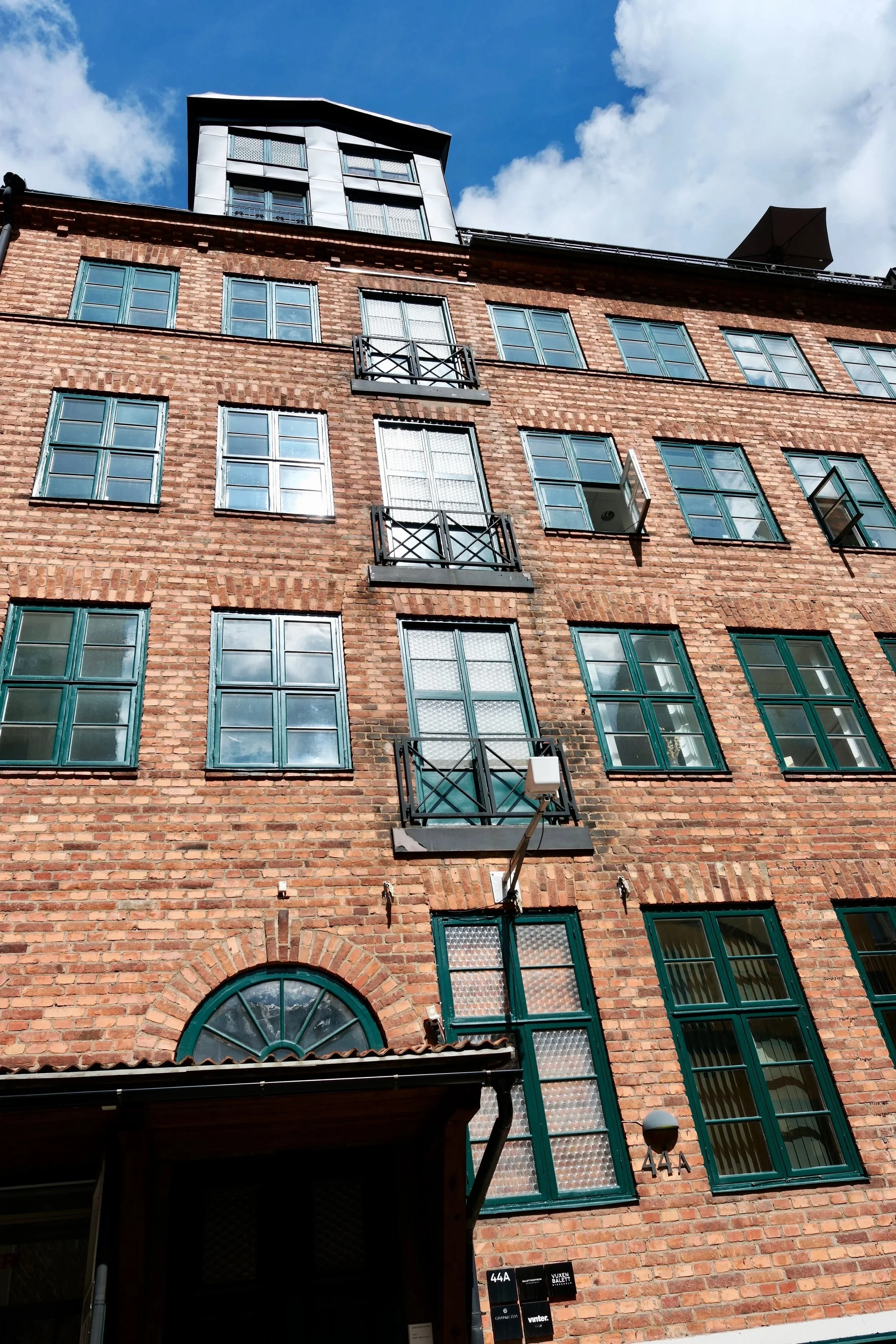 Bild av en röd tegelbyggnad med flera fönster, några öppna. Det finns en skylt med texten '44A' och en port under ett tak. Balettcentrum Stockholm