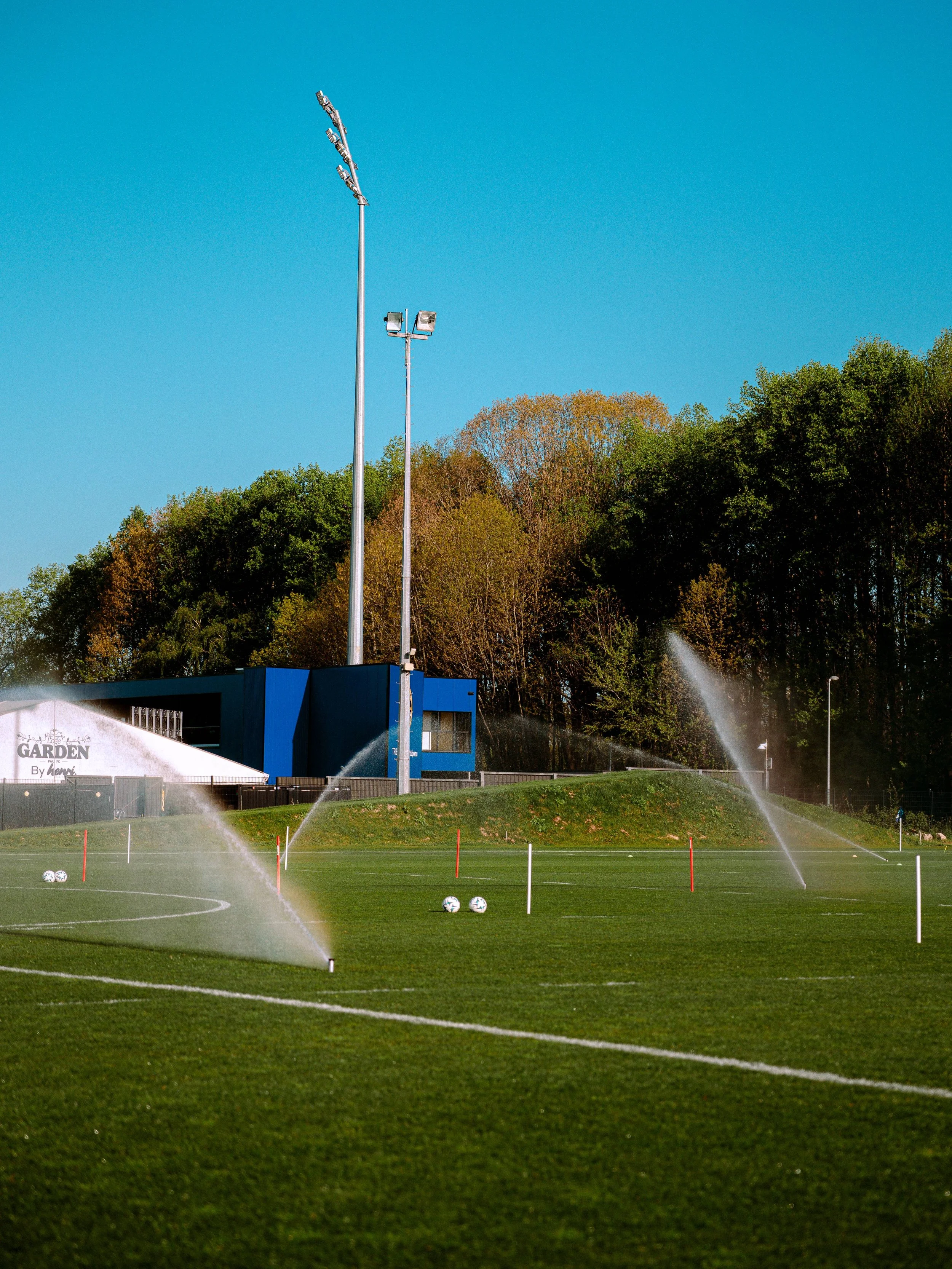 Un terrain de football avec des ballons et un arrosage automatique en marche, avec des lampadaires et un bâtiment bleu en arrière-plan, et des arbres verts sous un ciel bleu clair.