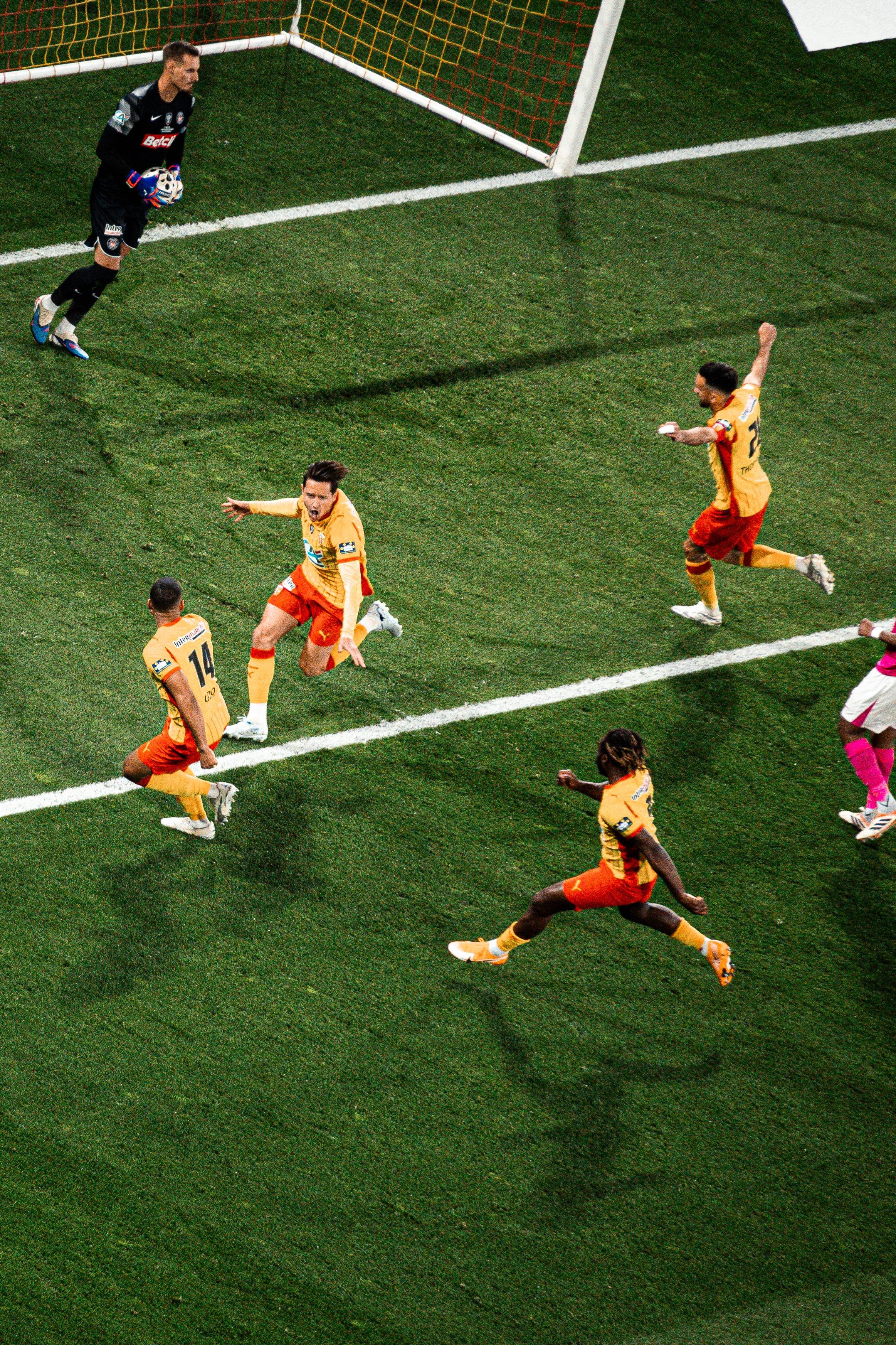 Une scène de football avec plusieurs joueurs en action près de la ligne de but, un gardien de but en position, et des joueurs en maillots jaunes et rouges en train de courir ou de sauter.