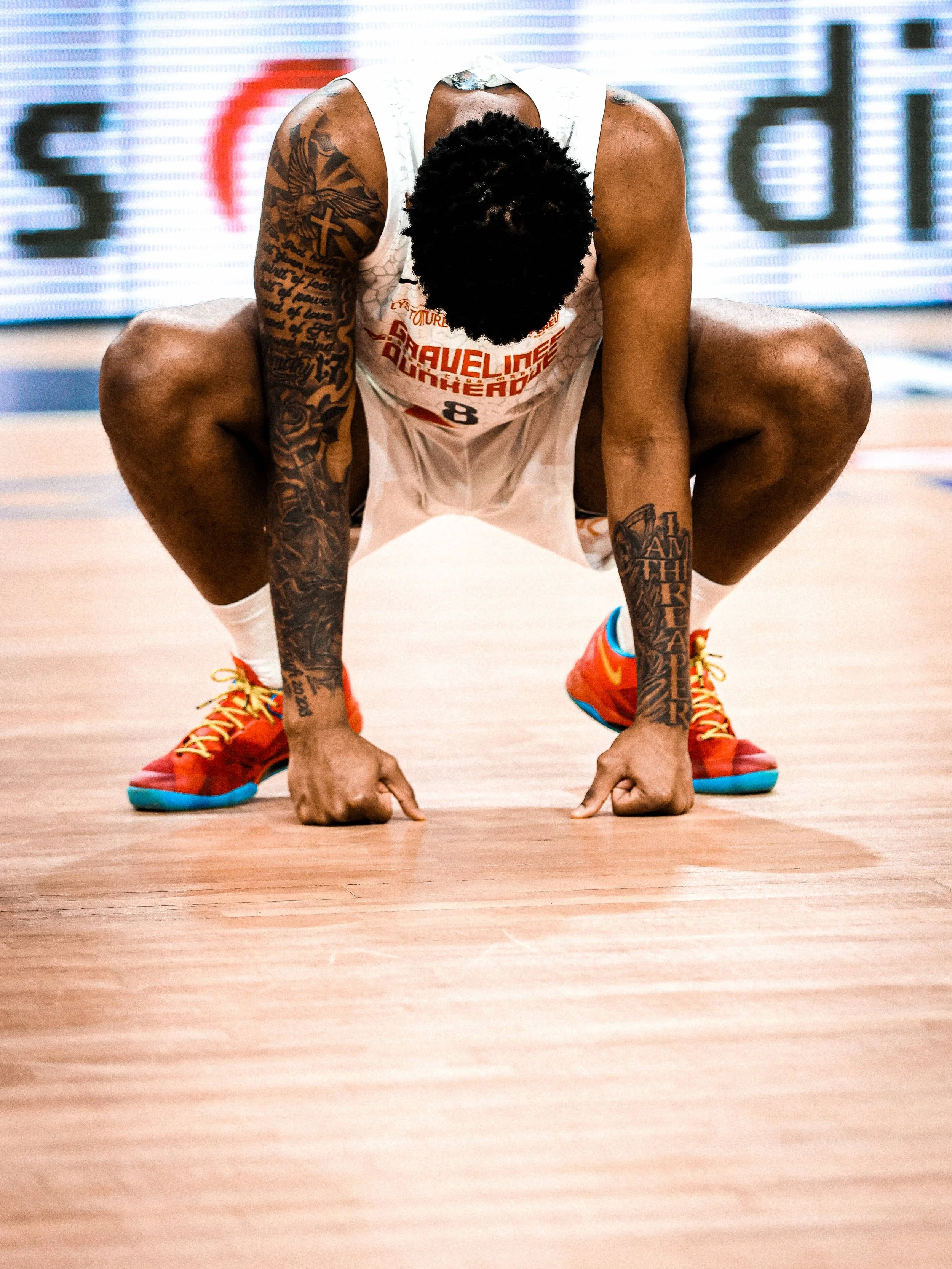 Un joueur de basketball en position accroupie sur le terrain, en train de préparer un mouvement, portant un maillot blanc, des tatouages visibles sur les bras, et des chaussures de sport orange et bleues.