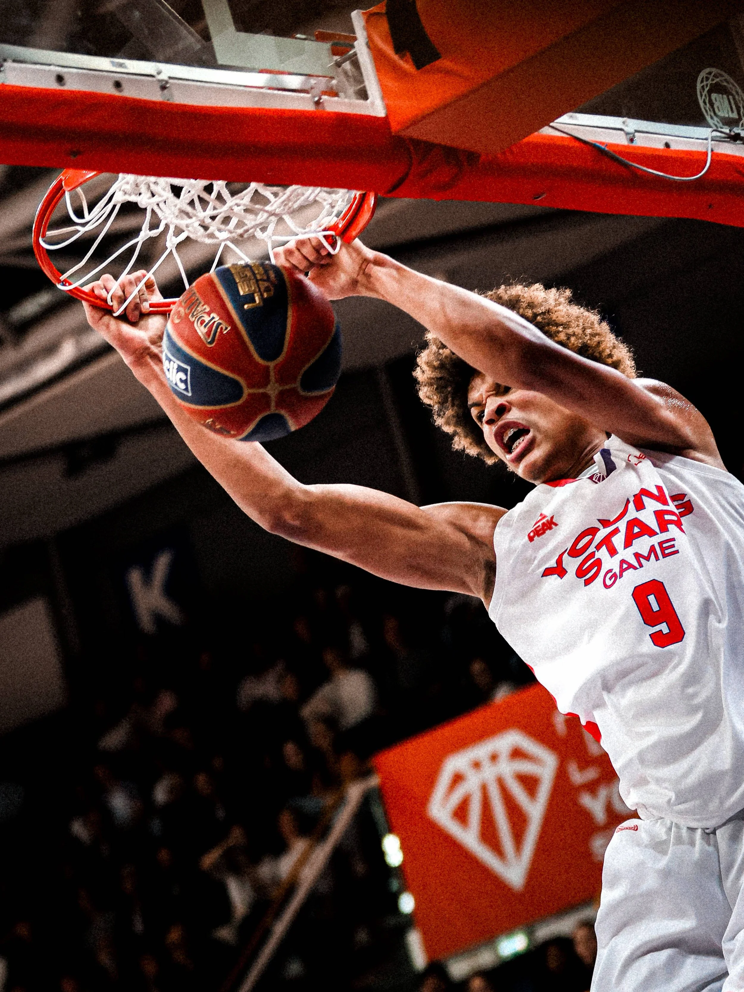 Un joueur de basketball en train de dunker au filet lors d'un match, portant un maillot blanc avec le logo de Yu-Gak Game et le numéro 9.