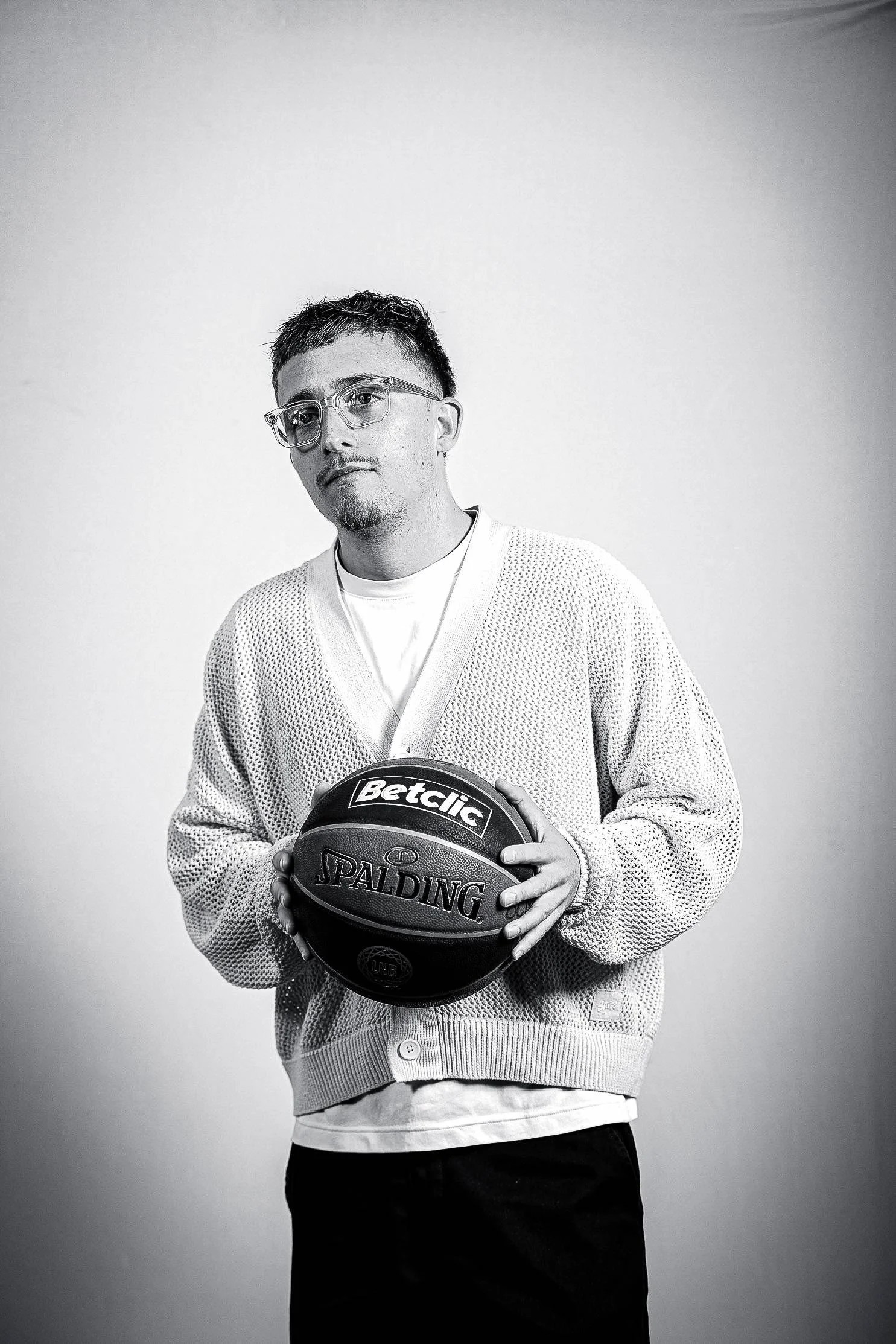 Un homme portant des lunettes, un pull en maille et un t-shirt blanc, tenant un ballon de basketball Spalding, posé devant un fond gris.
