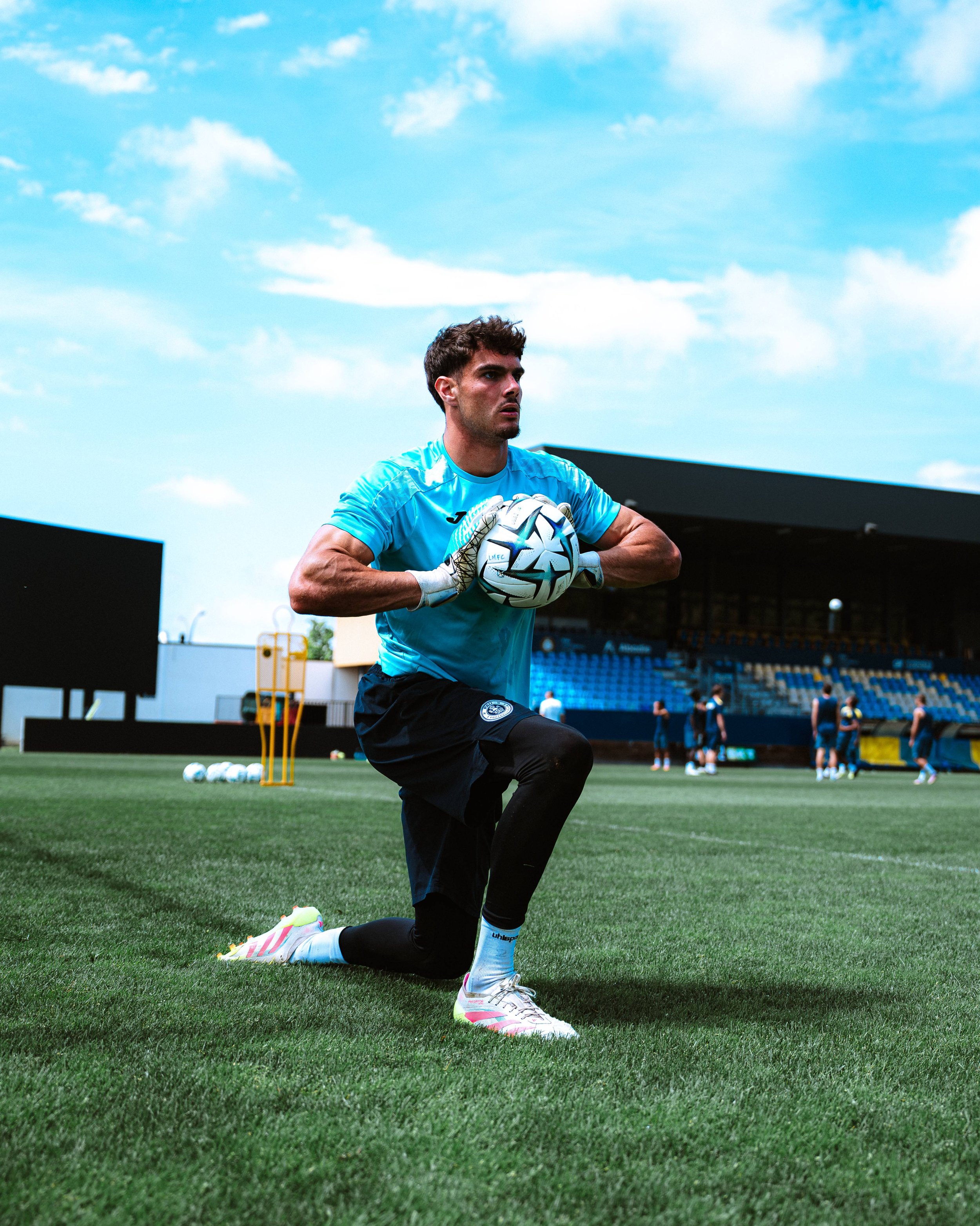 Un gardien de but en position sur un terrain de football, portant un maillot bleu, des gants et tenant un ballon blanc avec des motifs noirs. Il est à genoux sur l'herbe verte, avec d'autres joueurs en arrière-plan et un ciel bleu avec quelques nuage