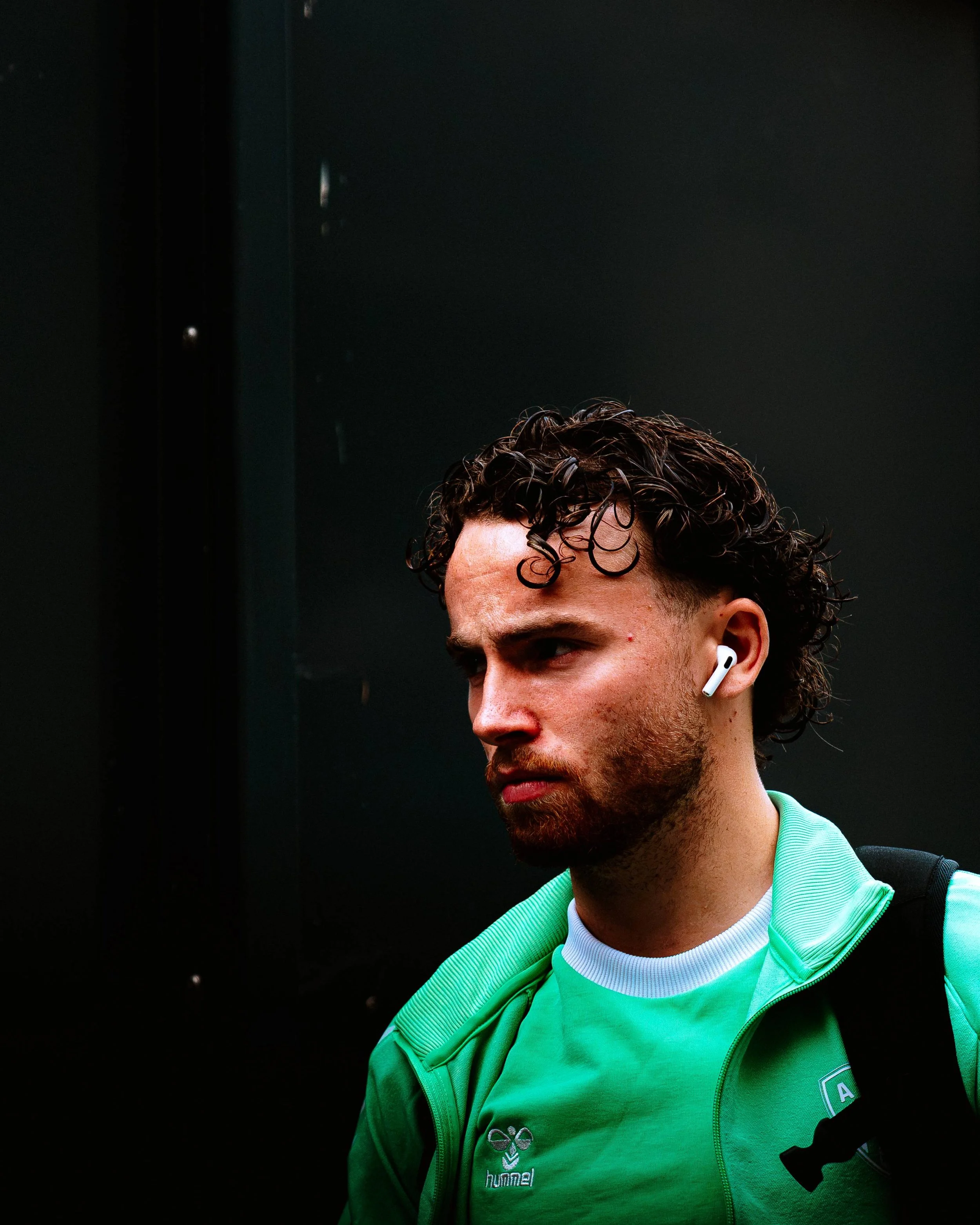 Jeune homme avec cheveux bouclés, portant un t-shirt blanc et une veste verte avec un logo Hummel, écouteurs sans fil et un sac à dos noir, dans un environnement sombre.