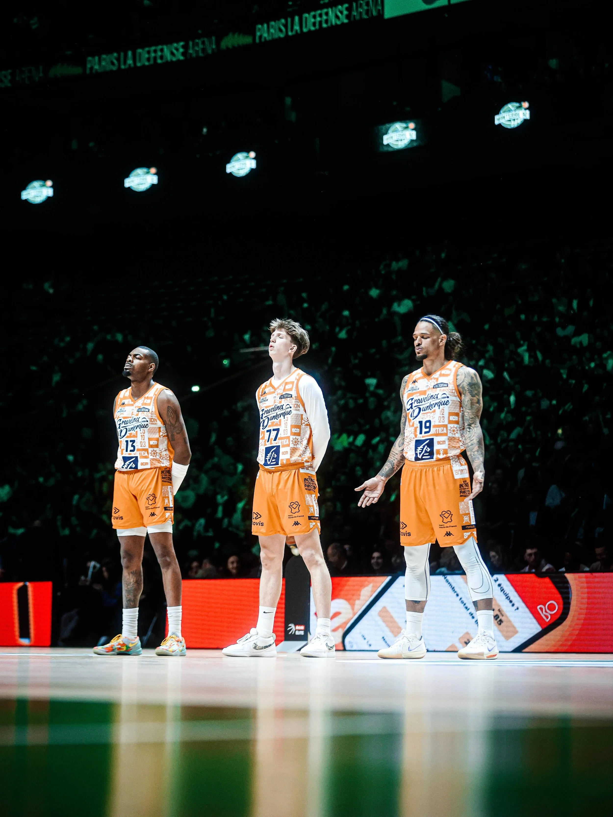 Trois joueurs de basketball en uniforme orange, debout sur le terrain lors d'un match, le public et les écrans en arrière-plan.