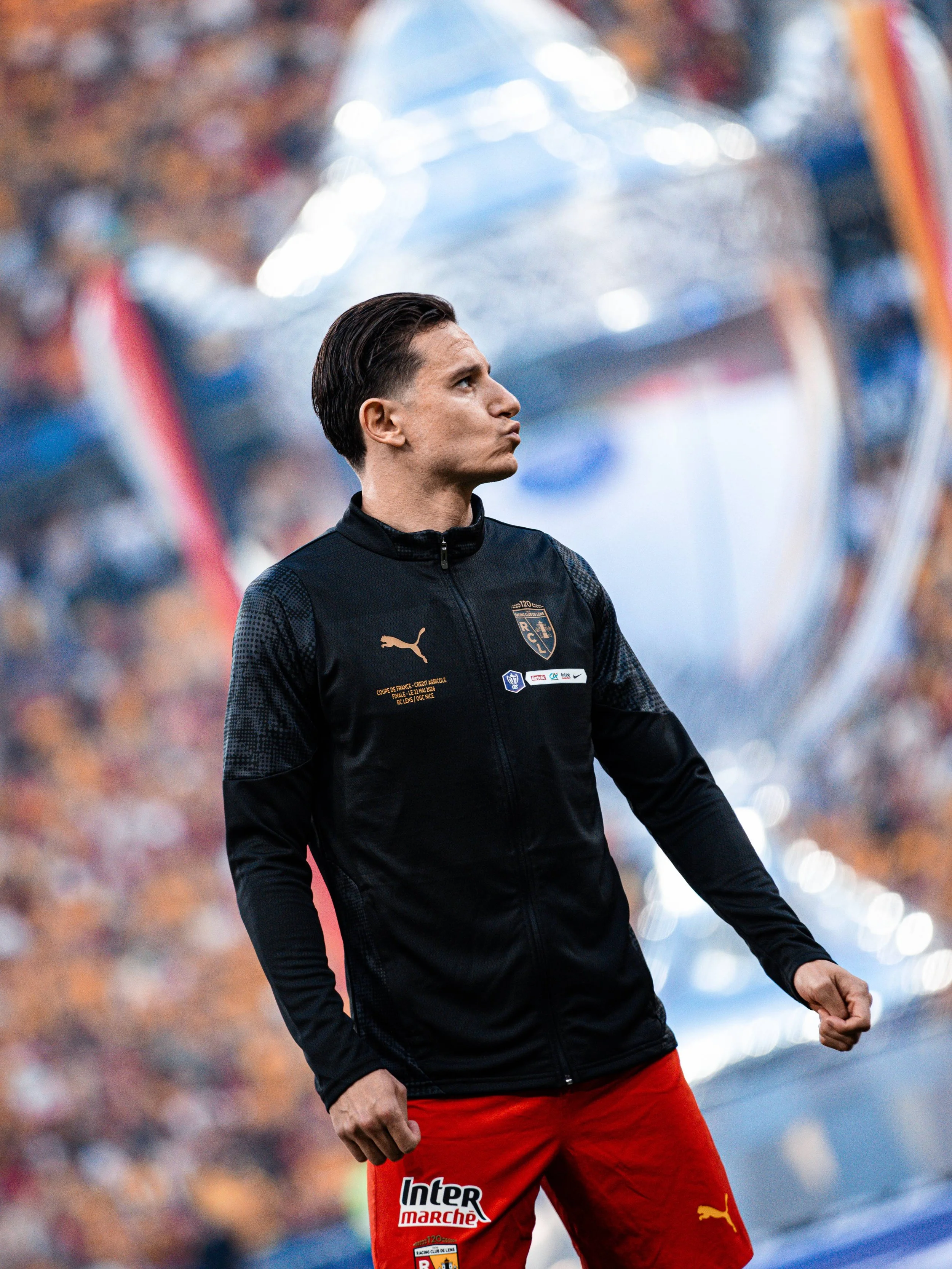 Un joueur de football en tenue de sport, portant un maillot noir avec des logo et des sponsor, et un short rouge, lors d'un match. Il fait un geste avec le poing fermé, l'ambiance est festive avec un public en arrière-plan.