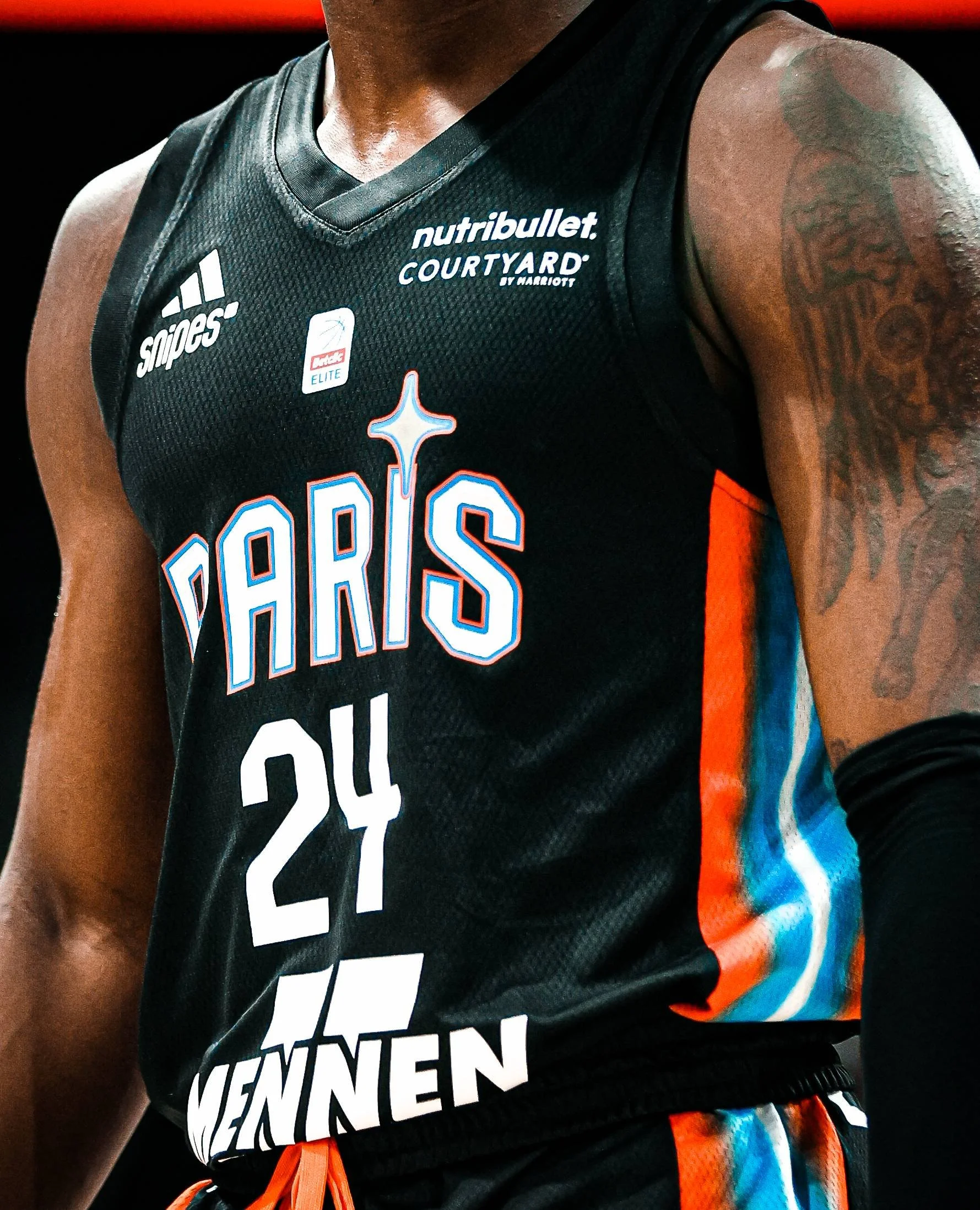 Un joueur de basket-ball portant un maillot noir avec des logos et une inscription en français, avec des rayures orange et bleue sur le côté du maillot.