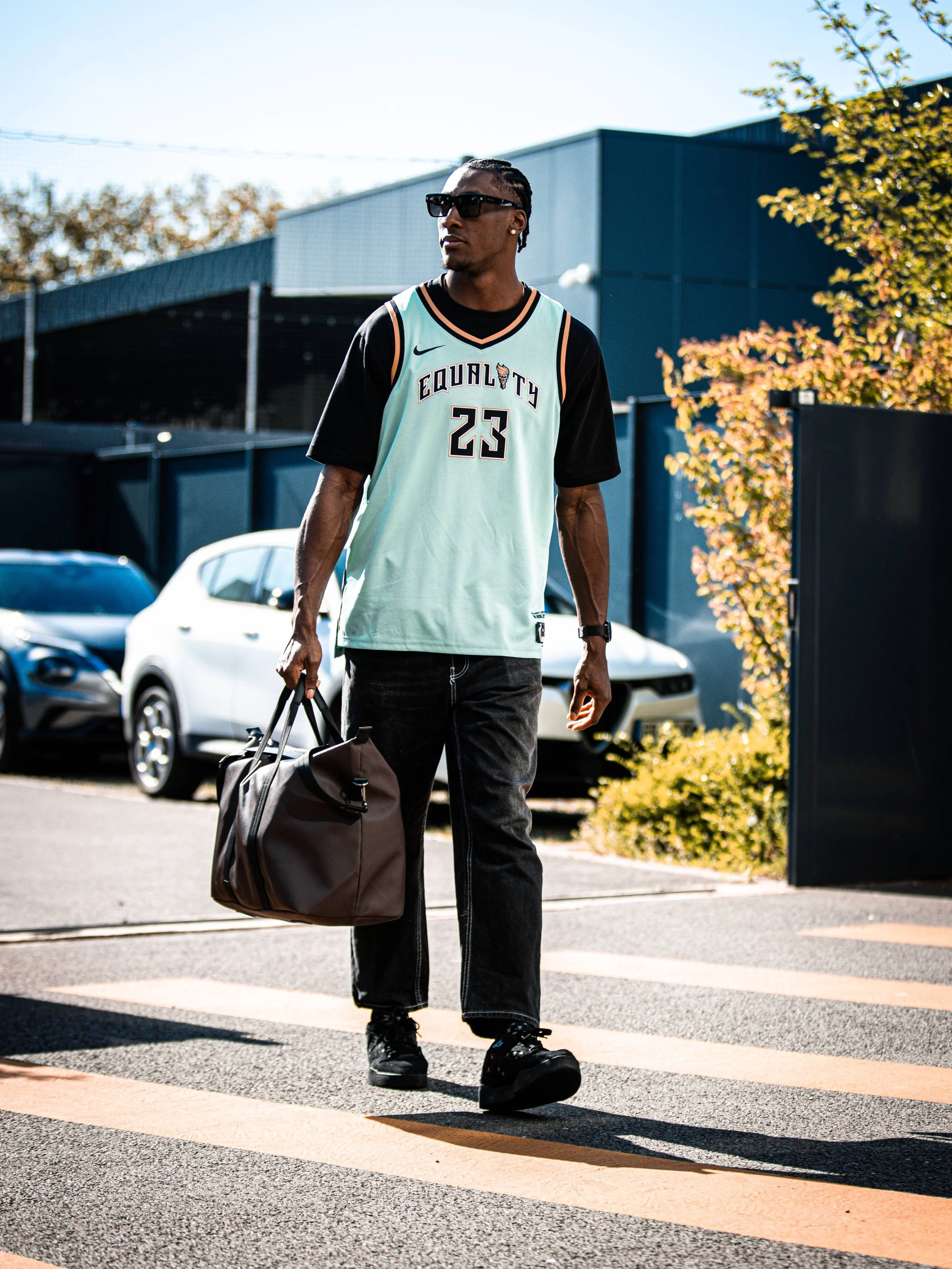 Un homme portant un maillot de basketball avec le numéro 23 et le nom 'Equality', des lunettes de soleil, marchant sur un passage pour piétons avec un sac de sport, sous un ciel ensoleillé.