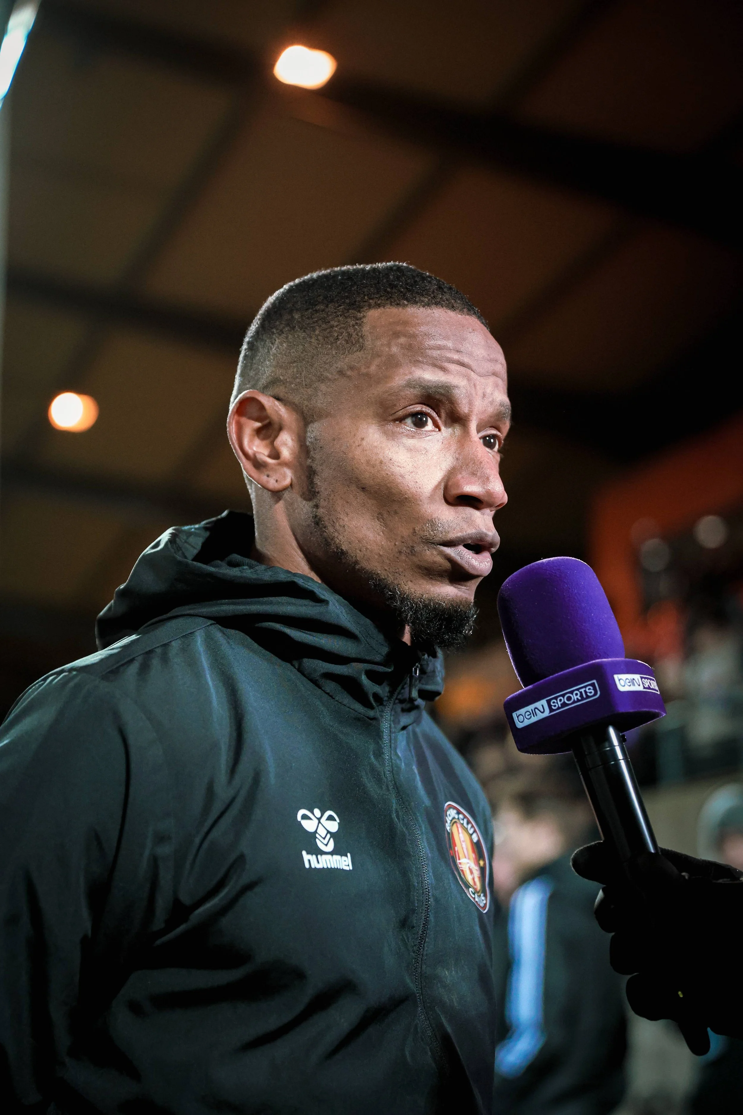 Homme noir parlant dans un micro violet portant une veste noire avec un logo de club sportif et un logo de la marque Hummel, probablement lors d'une interview ou d'une conférence de presse.