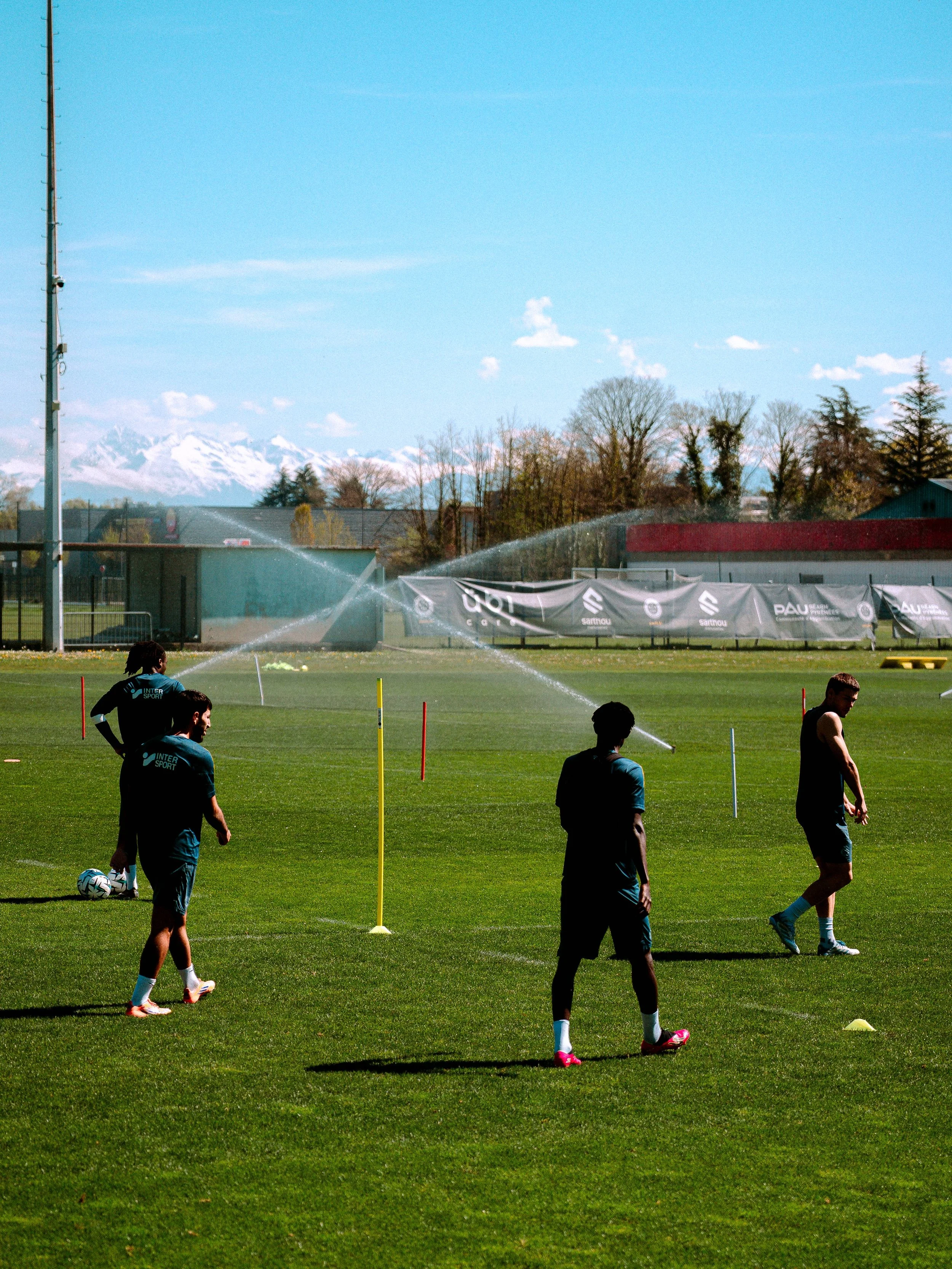 Des joueurs de football lors d'un entraînement sur un terrain vert, avec des arbres en arrière-plan et des montagnes enneigées au loin, sous un ciel bleu clair.