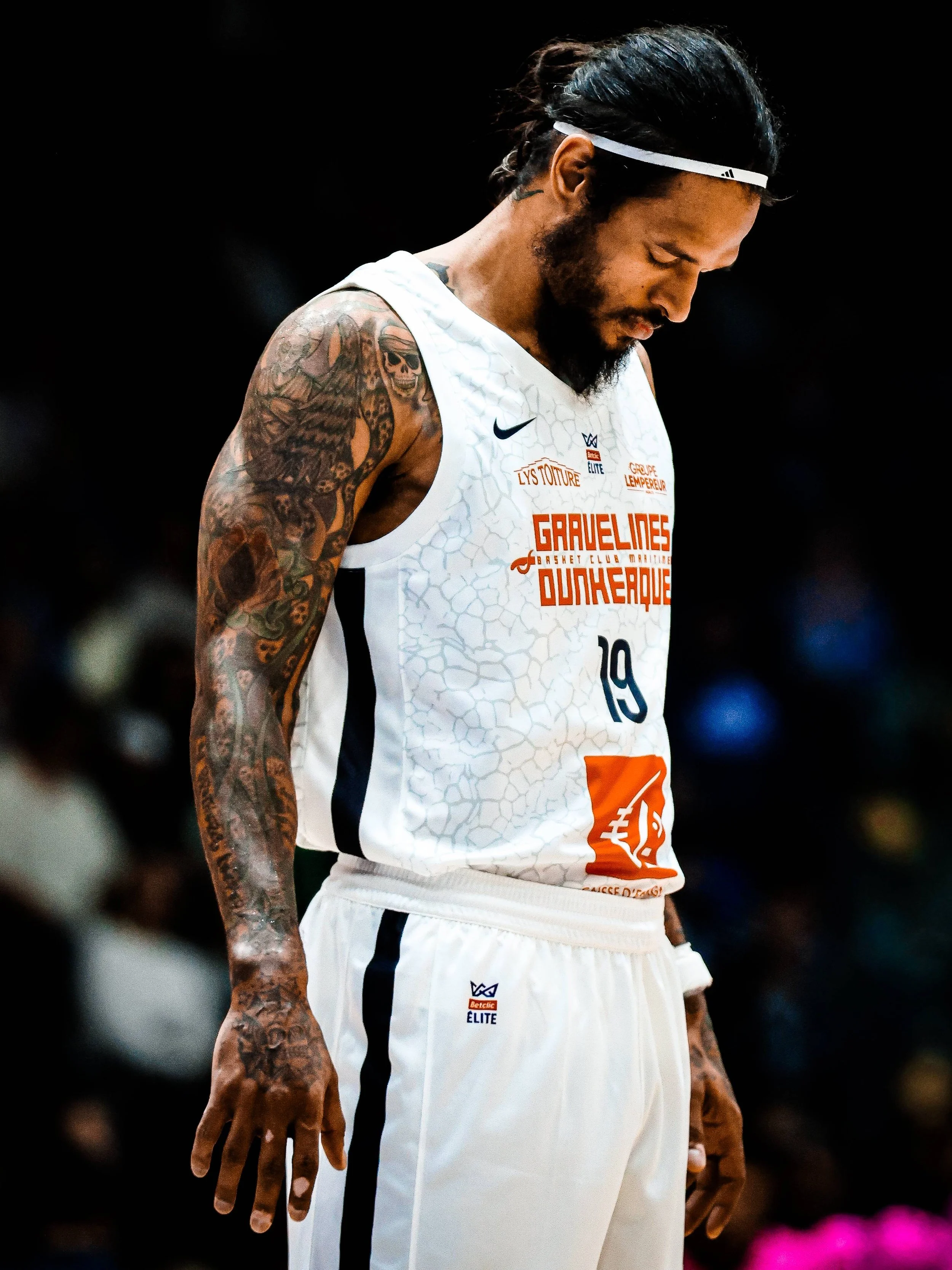 Un joueur de basket en costume blanc, portant un bandeau noir et une tatouage sur le bras, se tient le regard baissé, lors d'un match.
